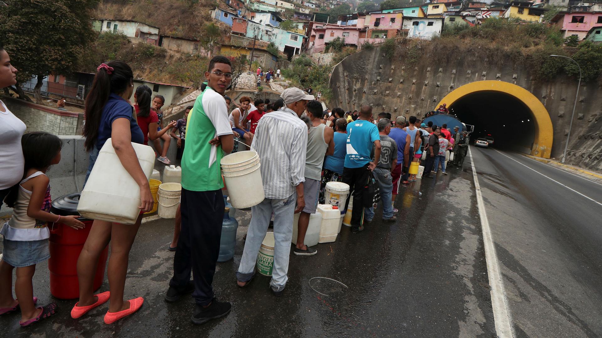 Dozens of people are shown standing in linewith buckets and other containers to collect water in Caracas.