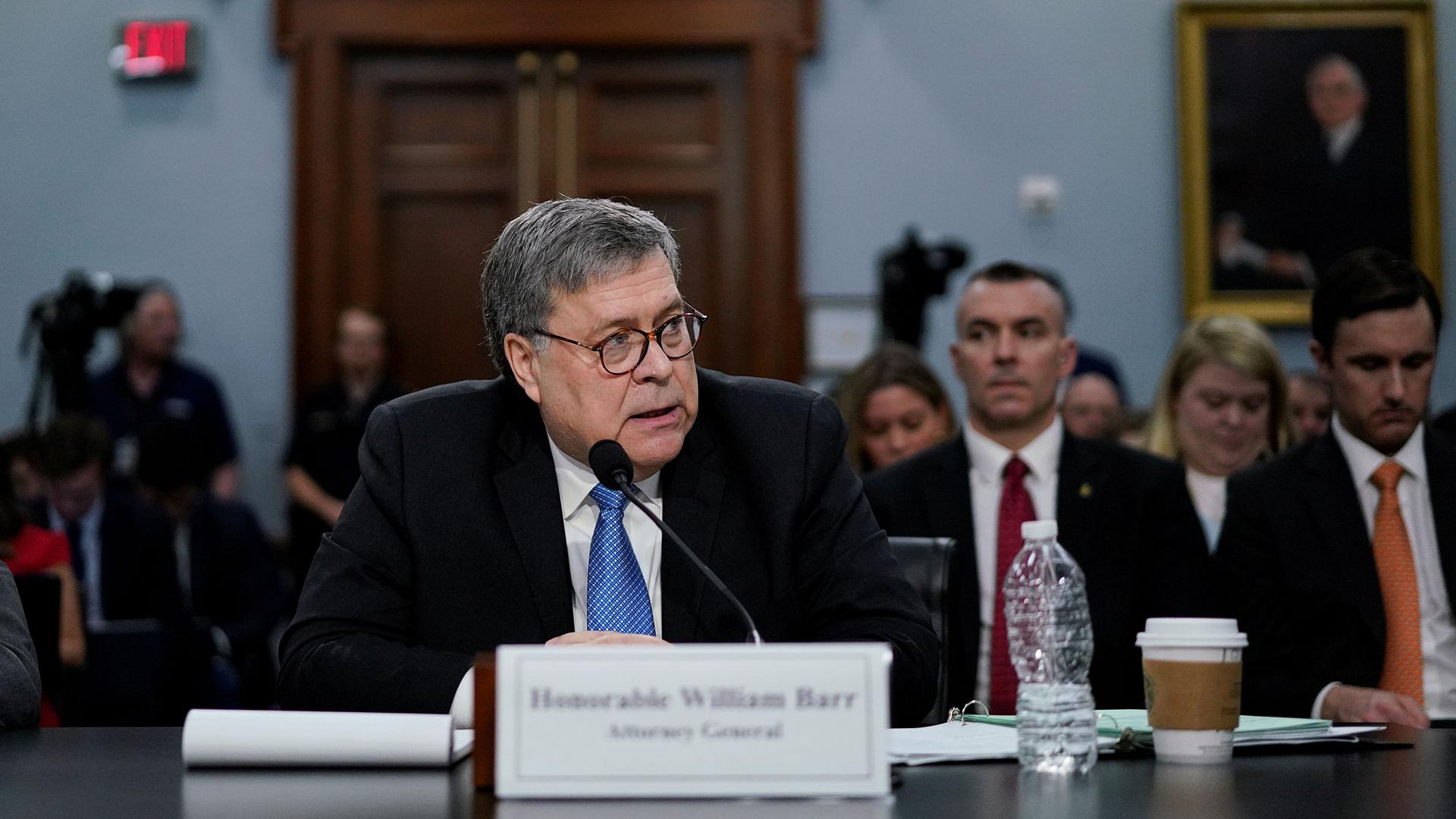 US Attorney General William Barr is shown sitting at a table behind a microphone and a sign with his name printed on it in soft focus.