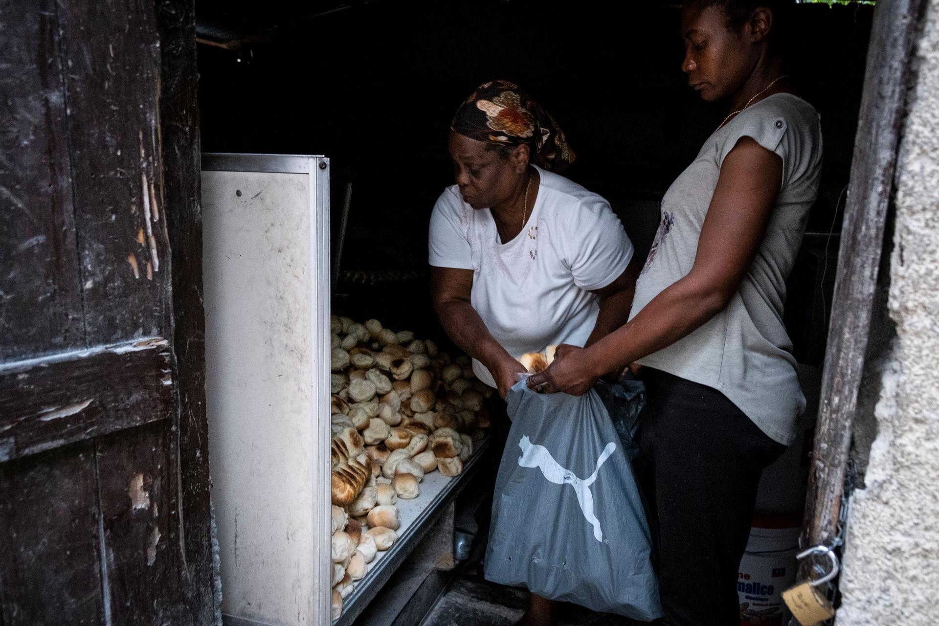 A woman is shown leaning over helping another woman load with biscuits into a plastic bag.
