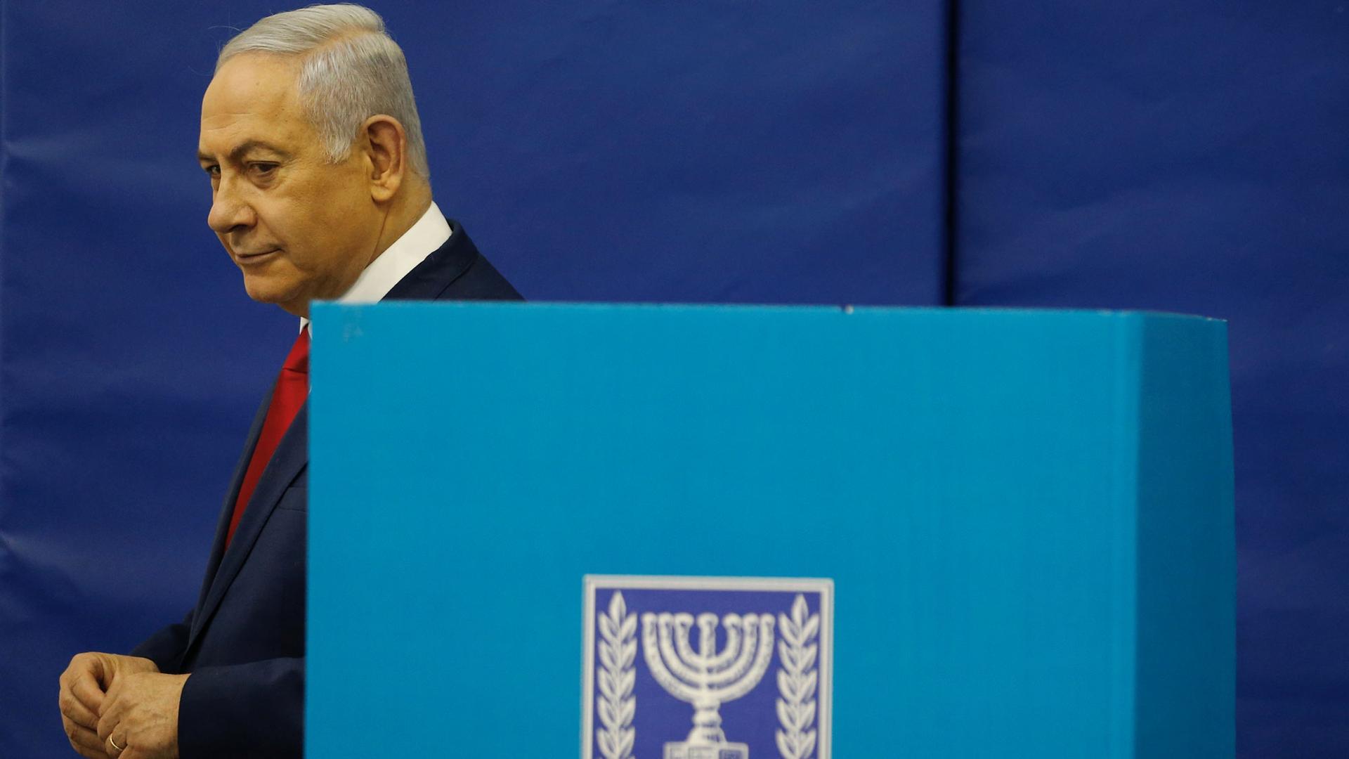 Israel’s Prime Minister Benjamin Netanyahu is shown wearing a blue suit and walking past a blue voting booth.