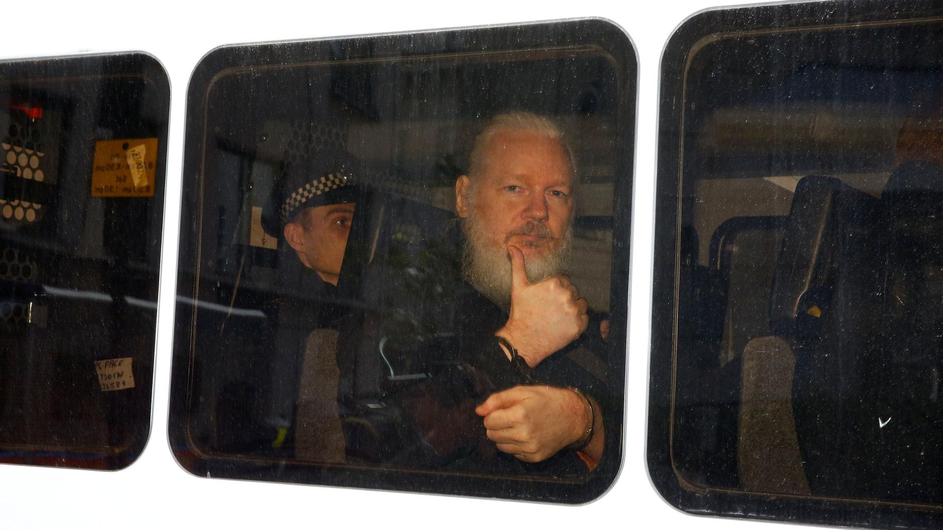WikiLeaks founder Julian Assange is seen with a long white beard, sitting and handcuffed in a police van.