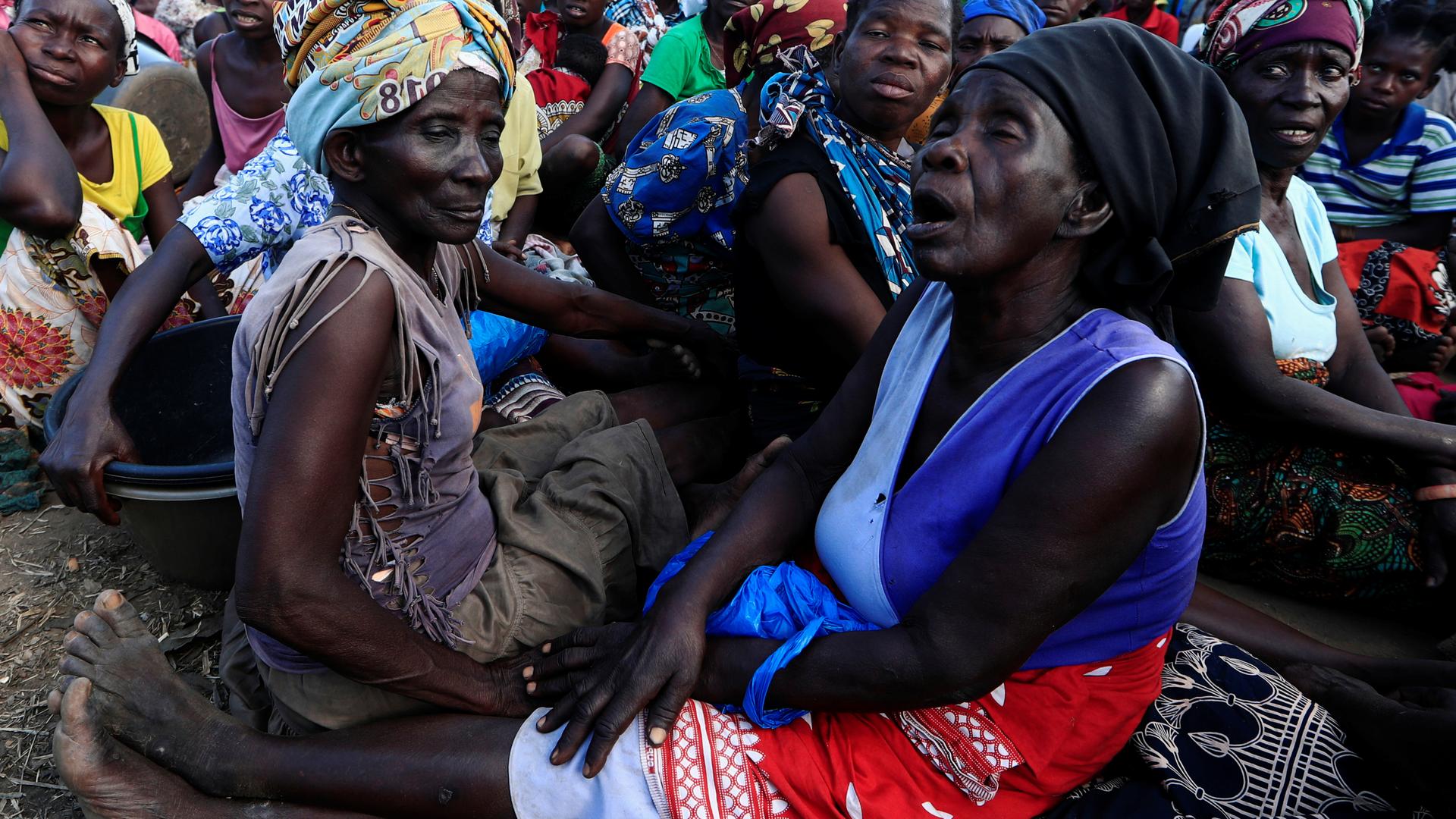 Women sit on the ground wearing colorful head wraps waiting for aid. 