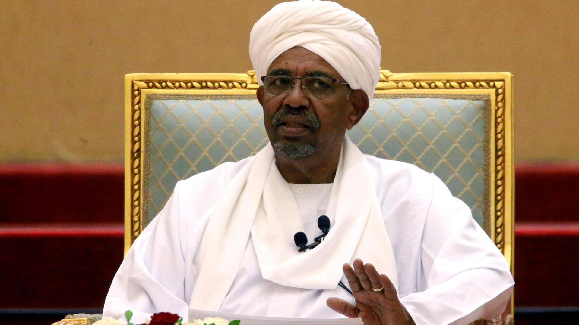 Omar al-Bashir is shown wearing all white while sitting in a chair with his left hand raised slightly. 