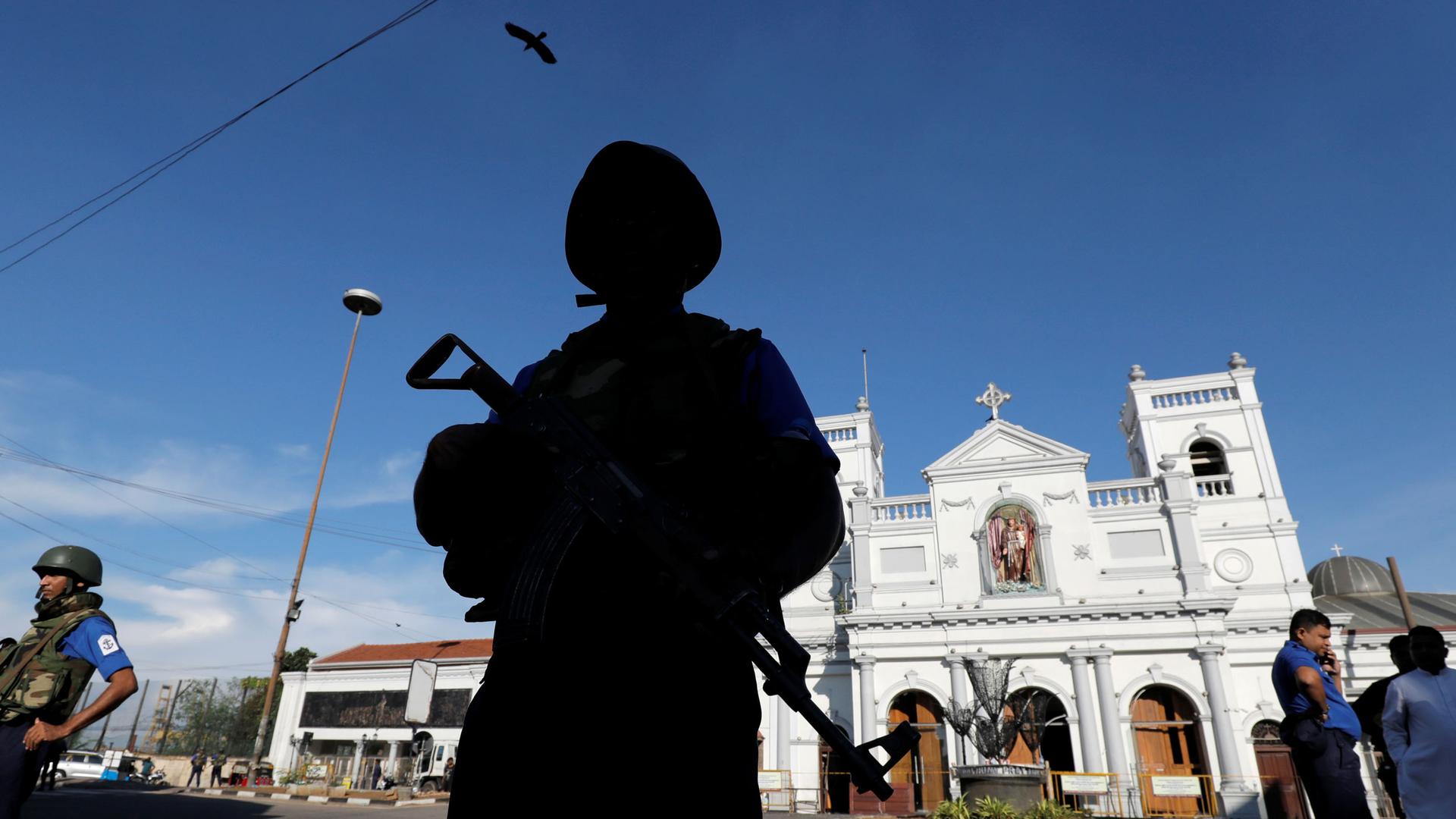 A security officer is shown standing in shadown and holding a gun with the white facade of St Anthony's shrine in the background.