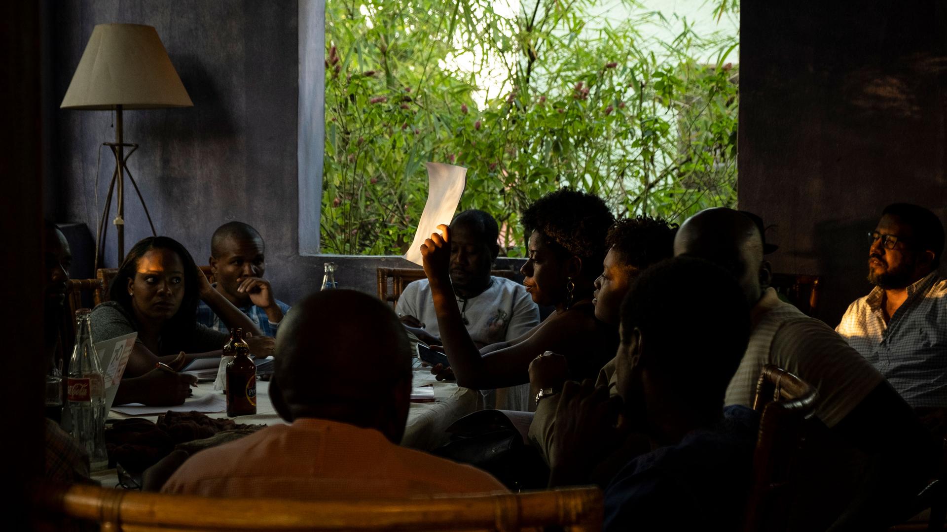 A group of young Haitians are shown sitting around a table in a room without glass in the windows.