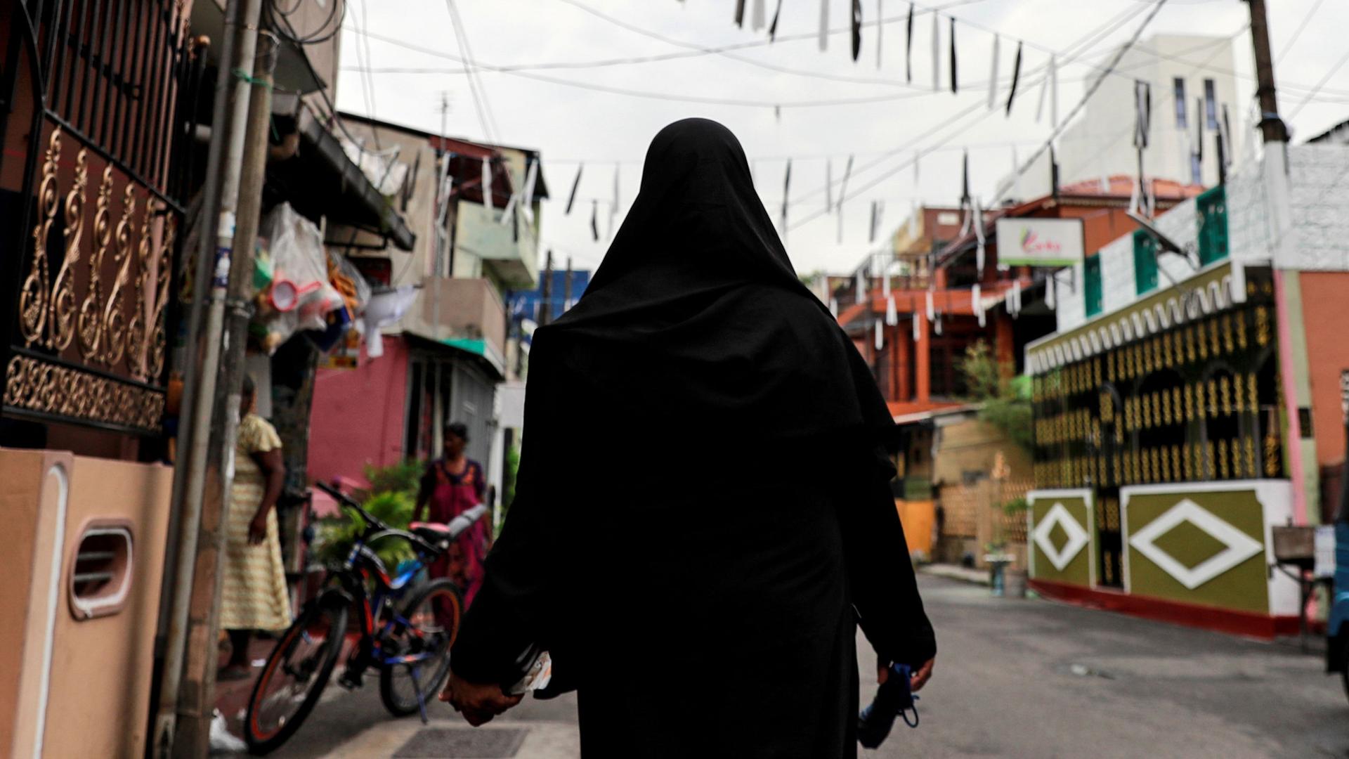 A woman is shown from behind wearing a black hijab walking through the middle of a street.
