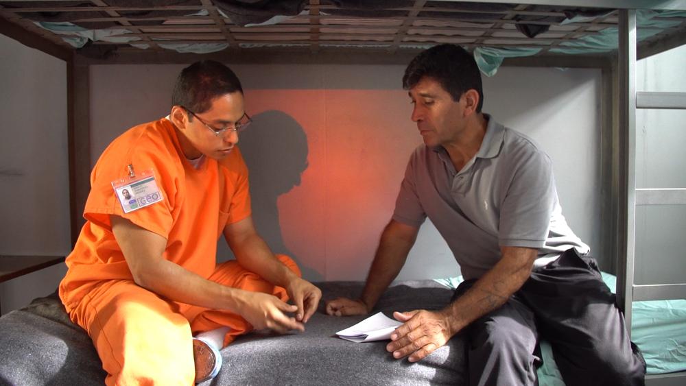 Two men sit on a prison bunk bed, one wearing an orange jumpsuit and the other in plain grey clothes. 
