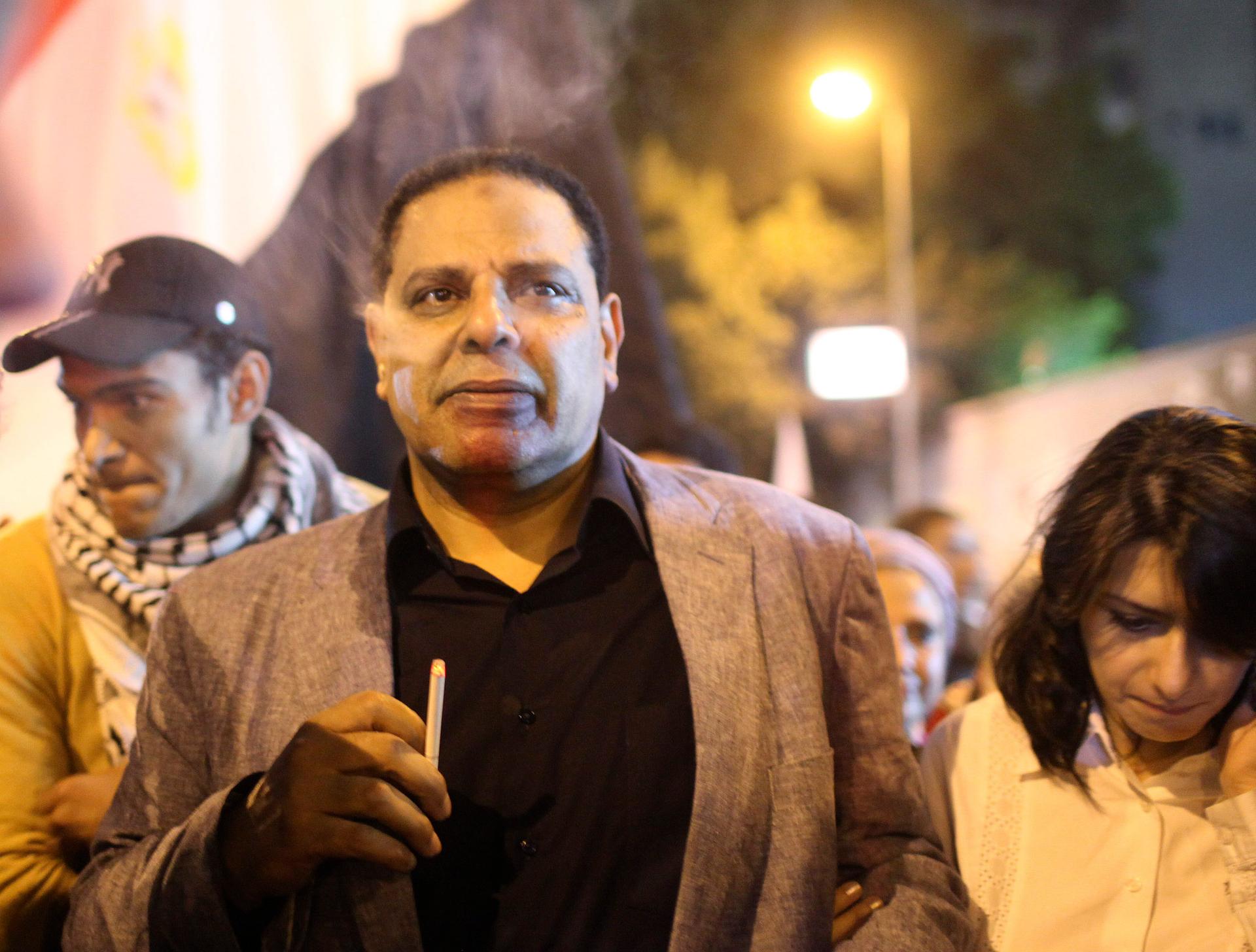 A man walks with a crowd at night with the Egyptian flag behind him. 