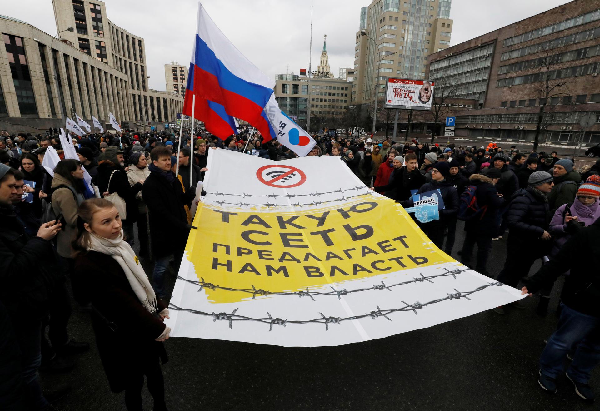 Protesters wa;l through the streets of a city carrying Russian flags and large signs written in Russian.