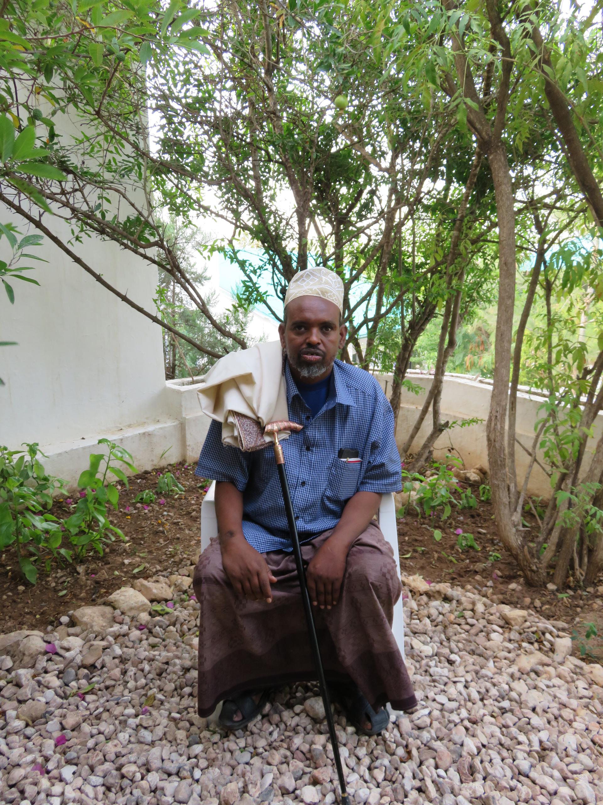 Somali man wears blue shirt and brown wrap with cream cap and holds a cane. 