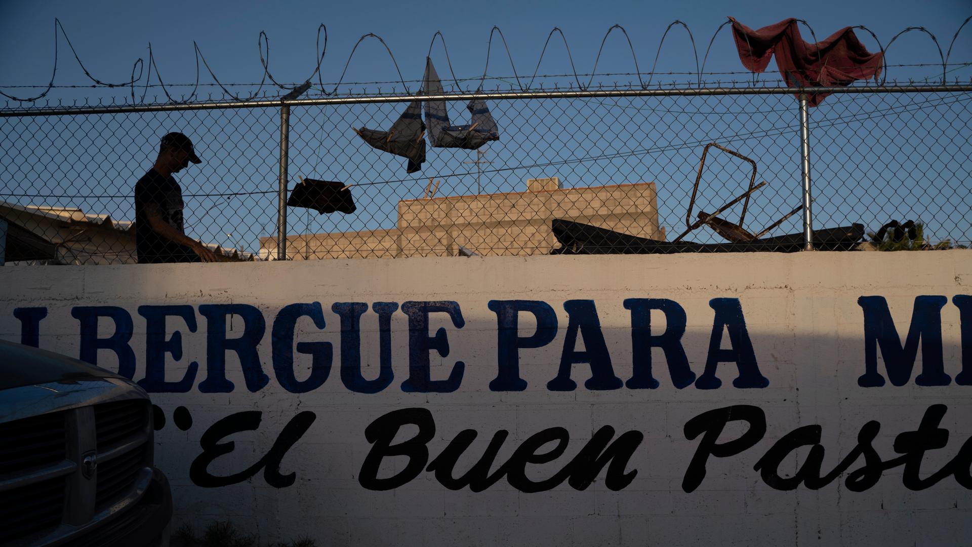 A man is silhouetted against a fence topped with razor wire. People have clothespinned clothing on the fence to dry. 