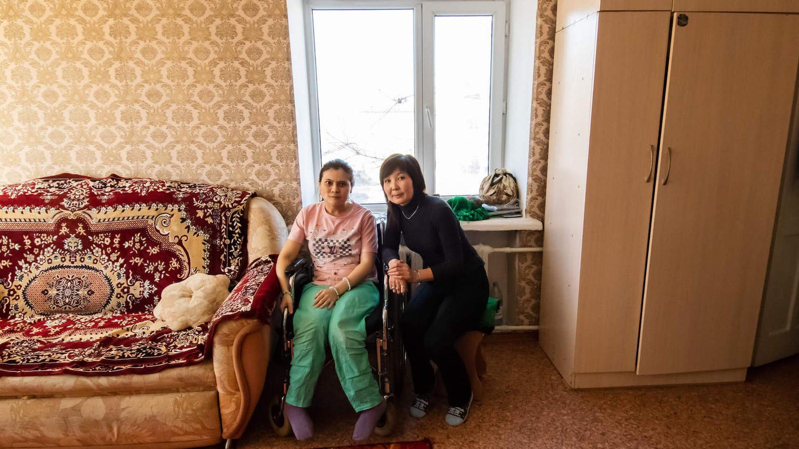 A young woman sits in a wheelchair next to her mom in the living room.