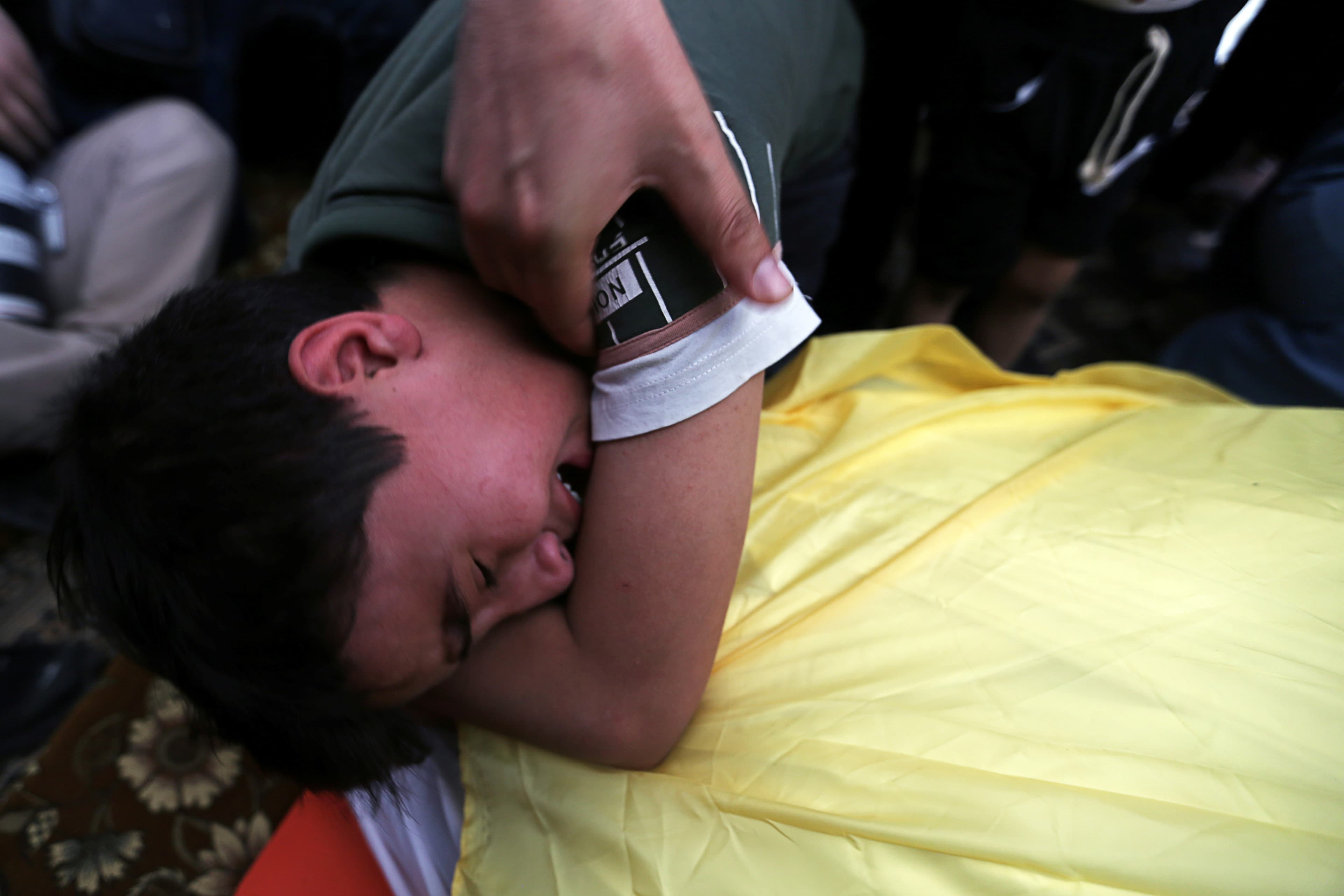 A boy sobs over the covered body of his father as a hand holds the boys shoulder.