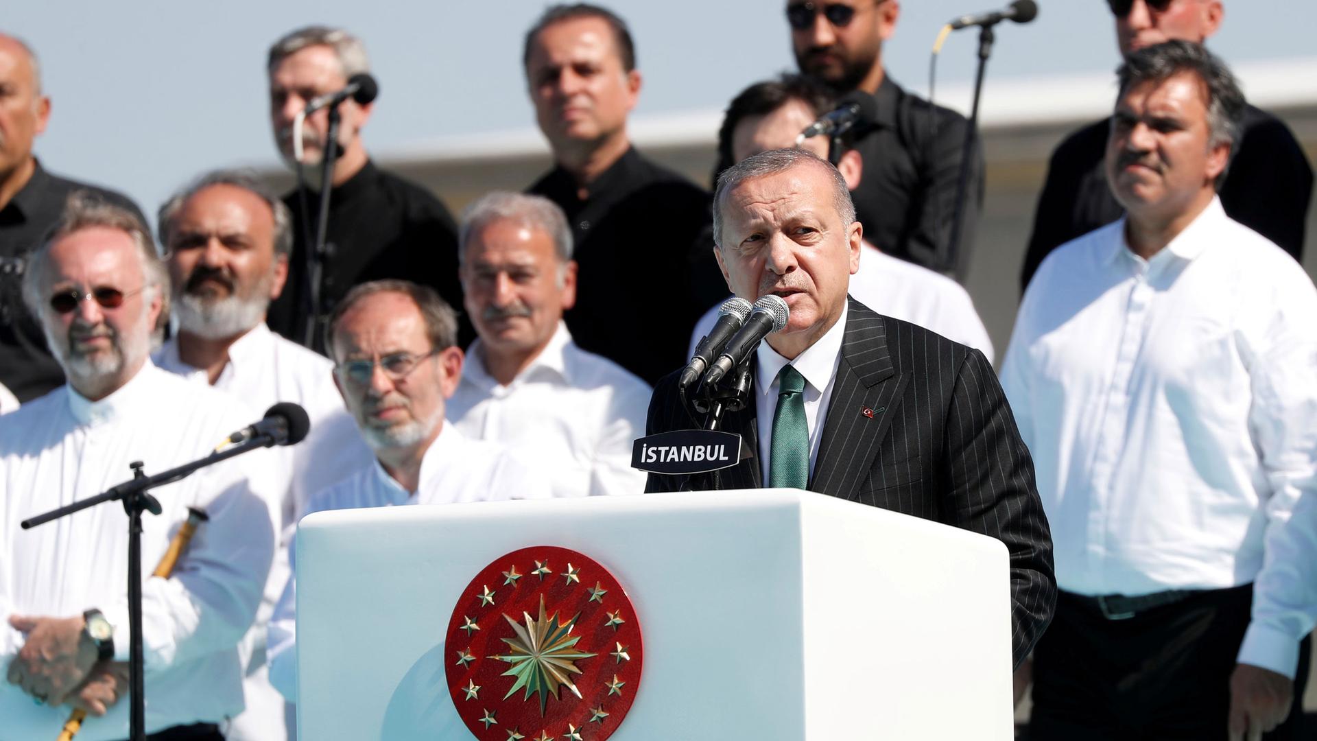 Turkish President Tayyip Erdoğan is shown making a speech behind a white podium with several stars on it.