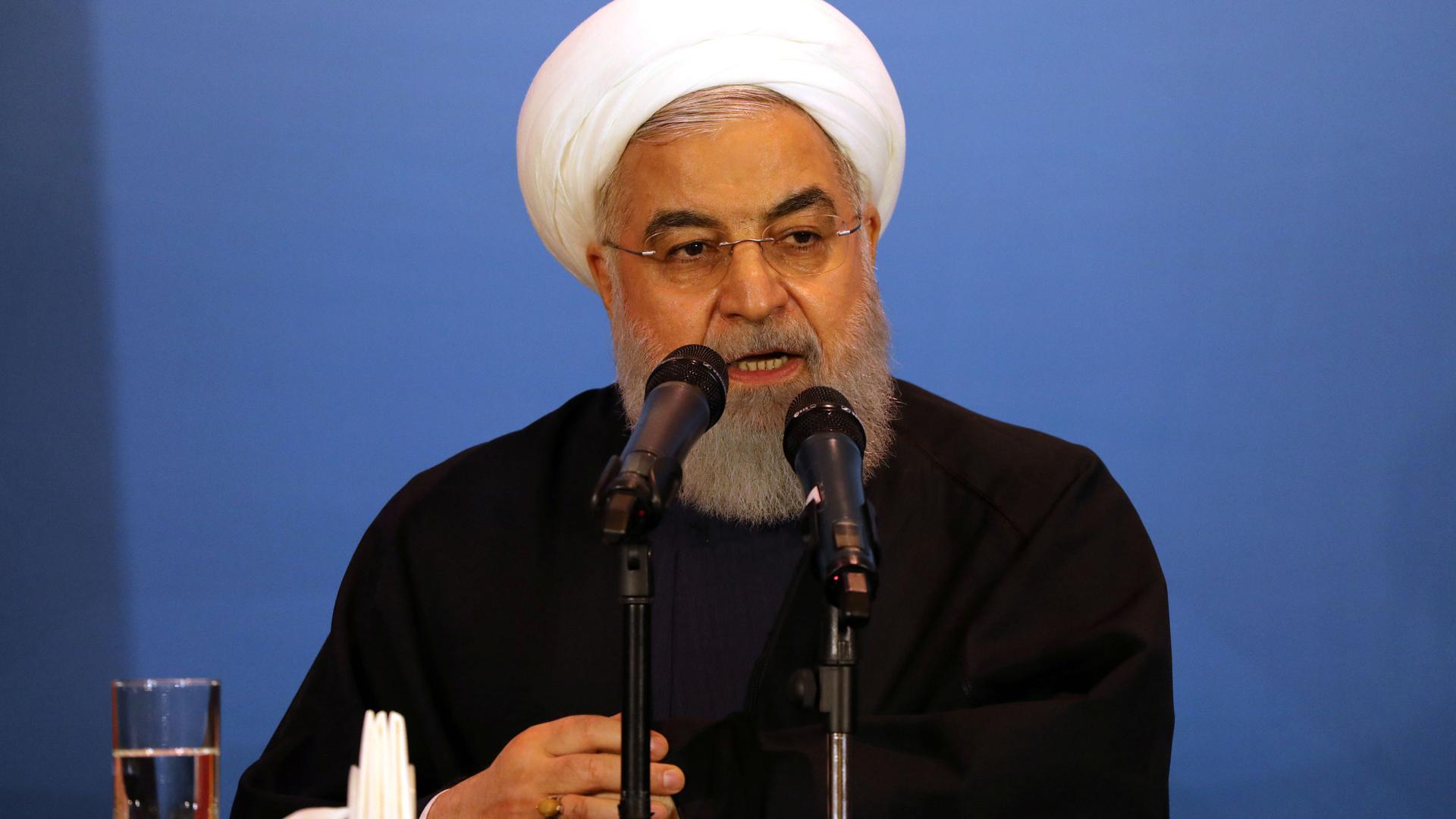 Iranian President Hassan Rouhani is shown sitting at a table with a glass of water speaking behind two microphones.