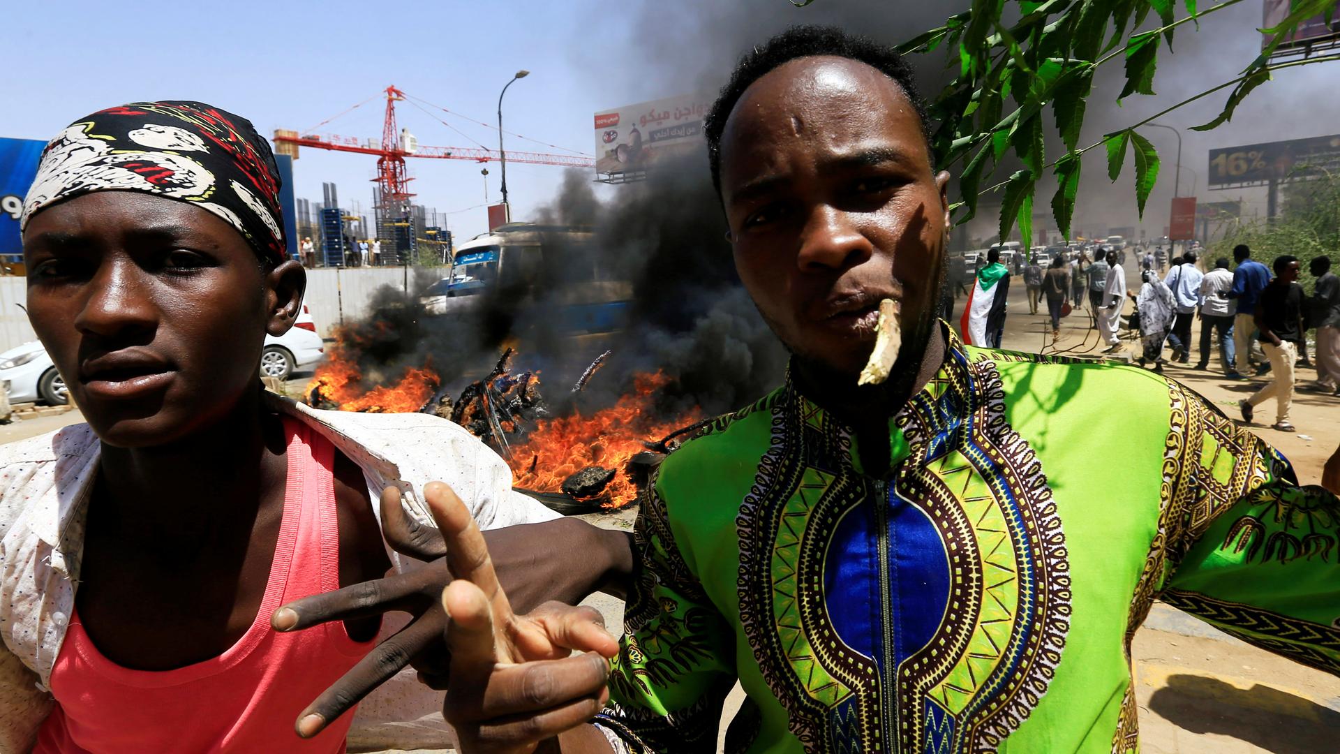 Two Sudanese men give a two-finger salute to the camera. Behind them a pile of tires is burning. 