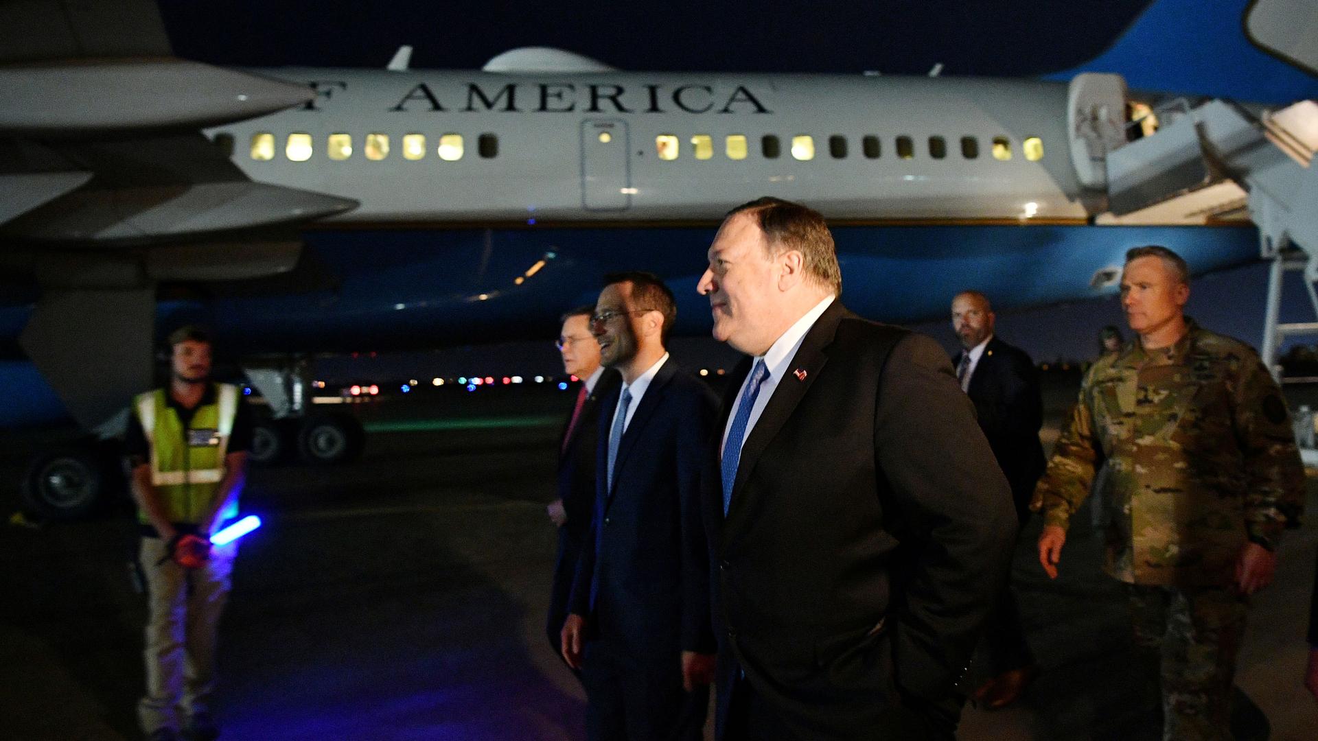 US Secretary of State Mike Pompeo is shown wearing a dark suit and blue tie while walking on the tarmac in Iraq with a State Dept. airplane in the background.