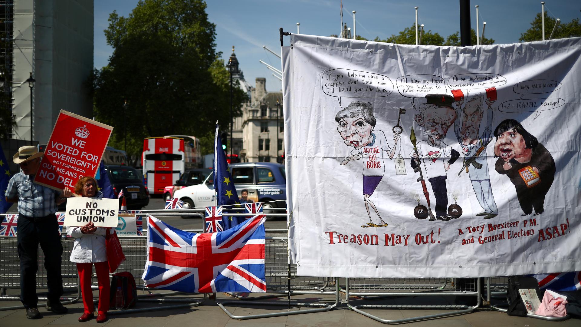 A man and a woman with pro-Brexit signs stand next to a banner of political caricatures. 
