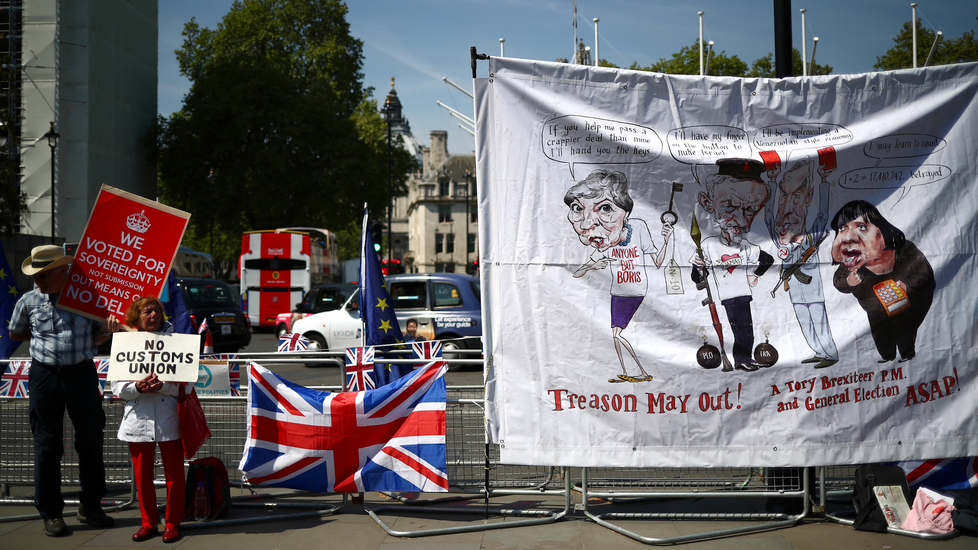 A man and a woman with pro-Brexit signs stand next to a banner of political caricatures.