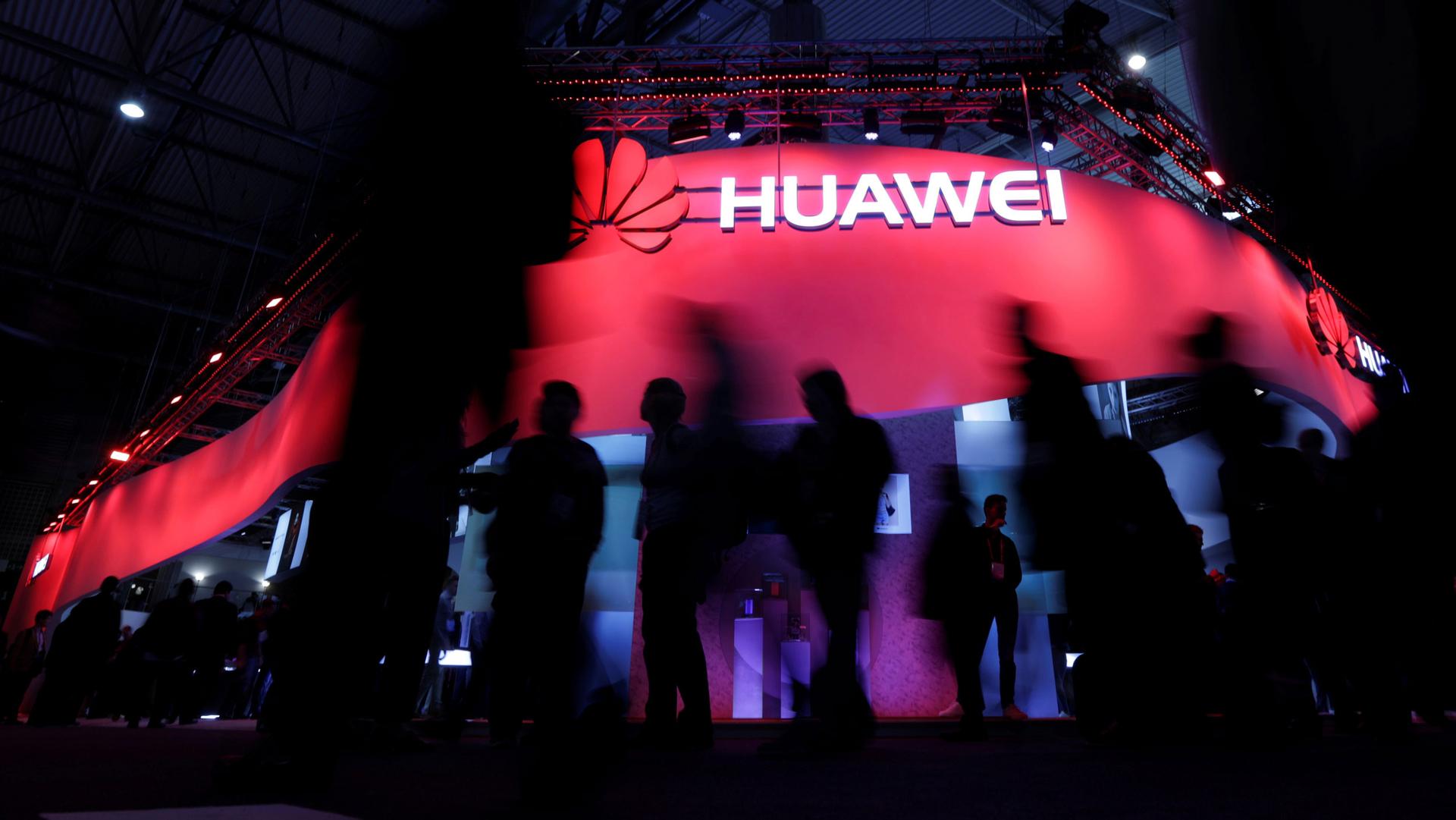 Several people in shadow are shown walking by a red illuminated sign for Huawei.