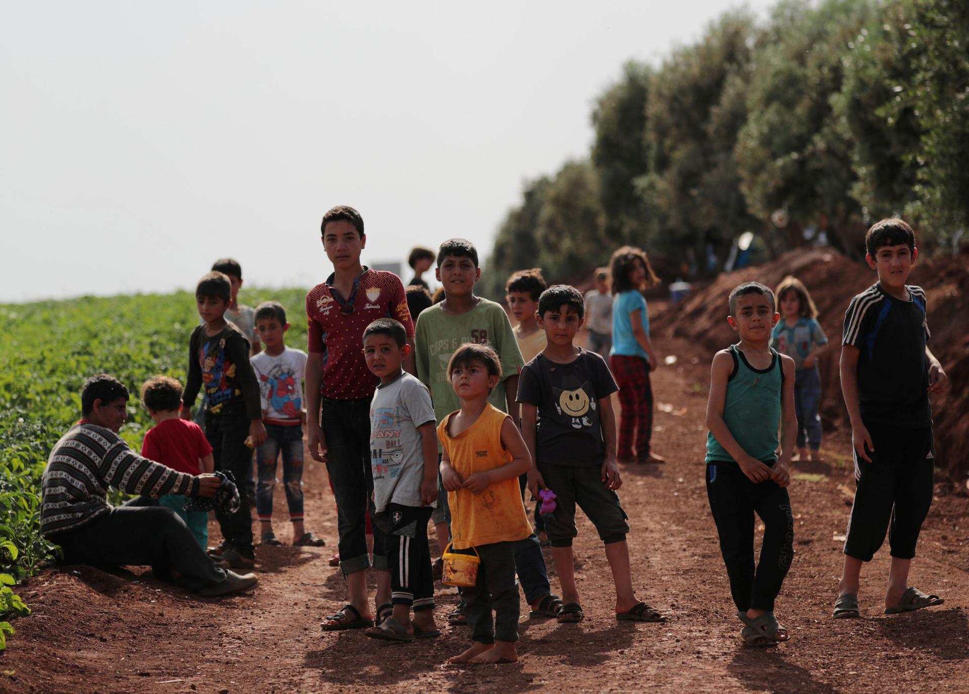 A group of more than a dozen children of varying age are shown standing in a dirt road next to a field.