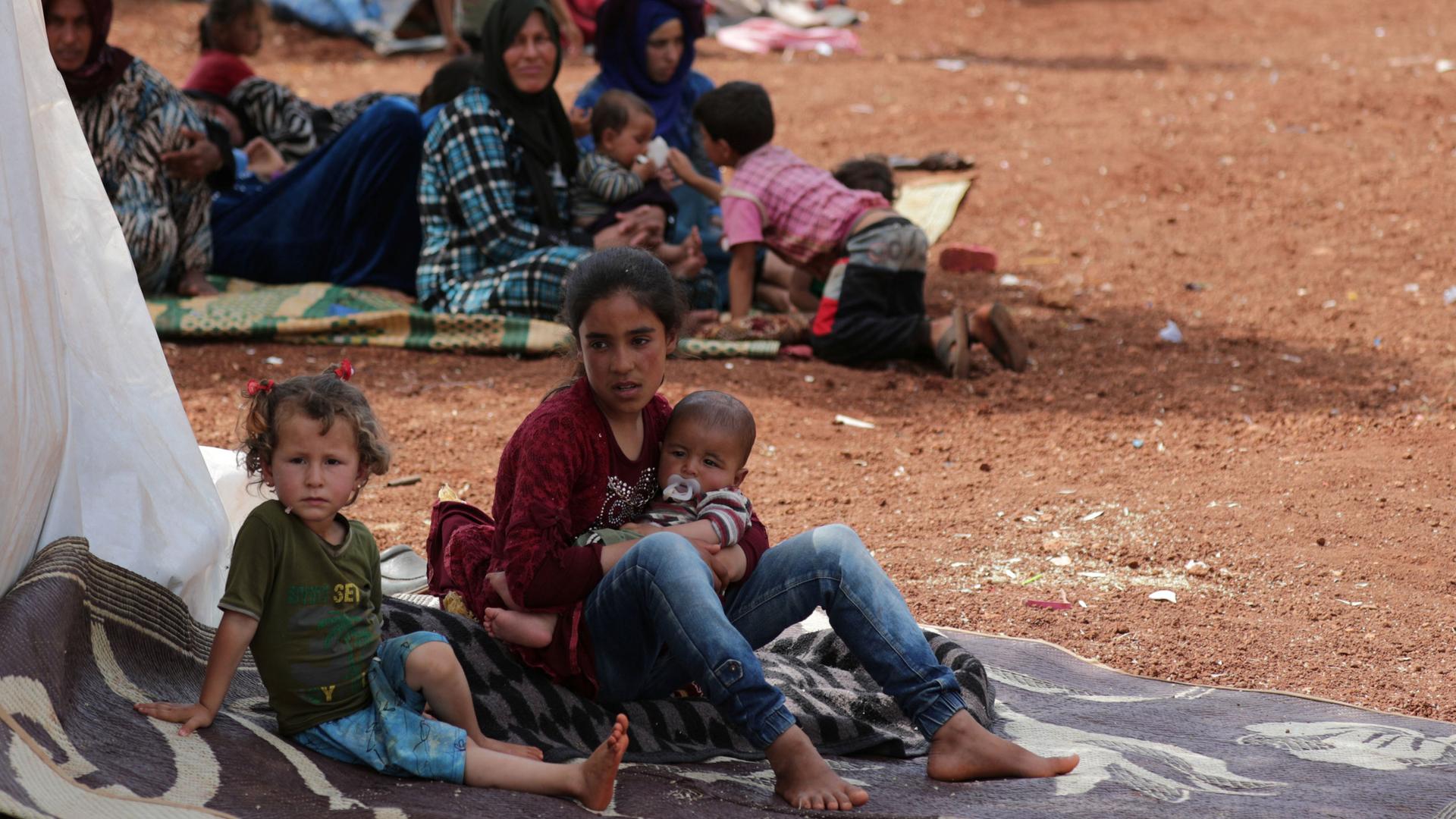 Two displaced Syrian girls is shown sitting on a mat and one holding a baby.