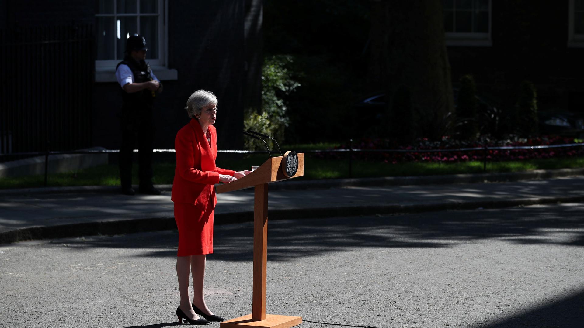 British Prime Minister Theresa May is shown standing at a wooden podium and wearing a red suit.