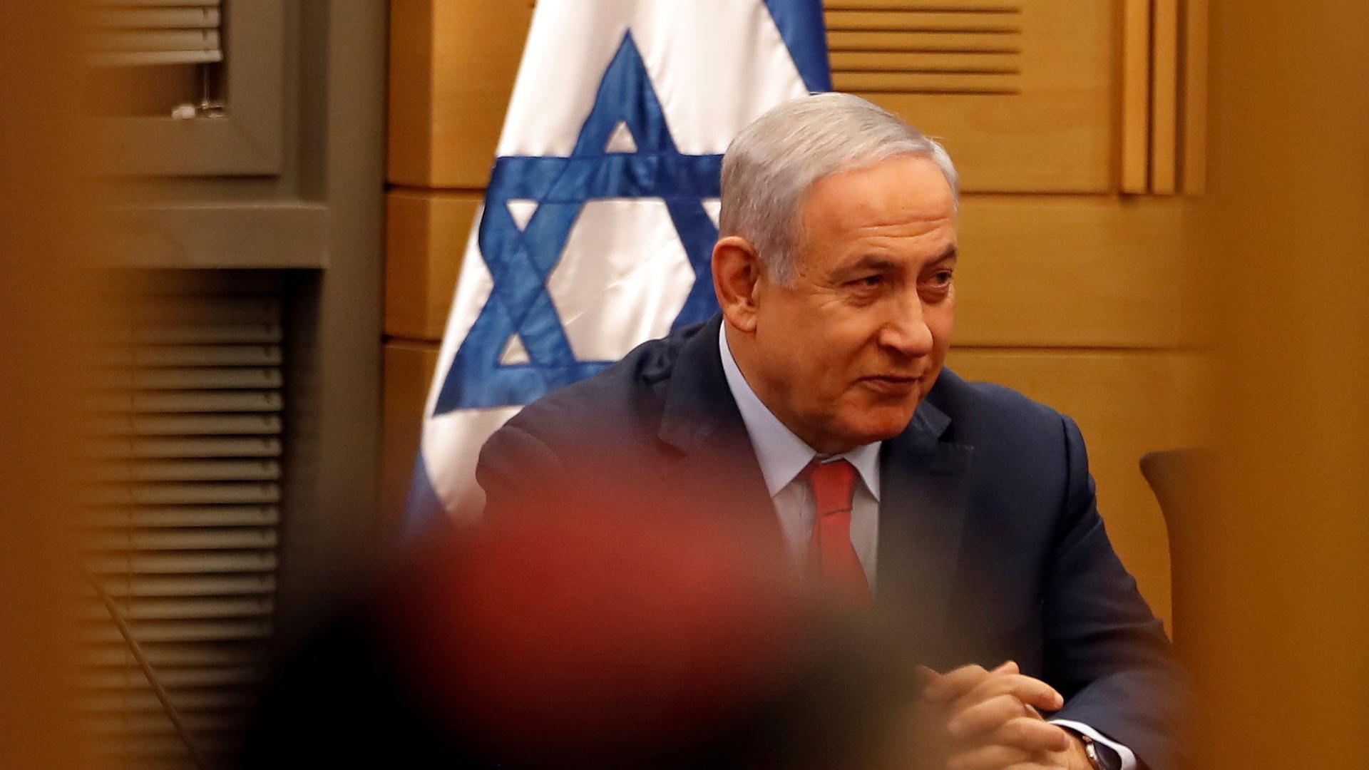 Israeli Prime Minister Benjamin Netanyahu is shown sitting with his hands folded and an Israeli flag behind him.