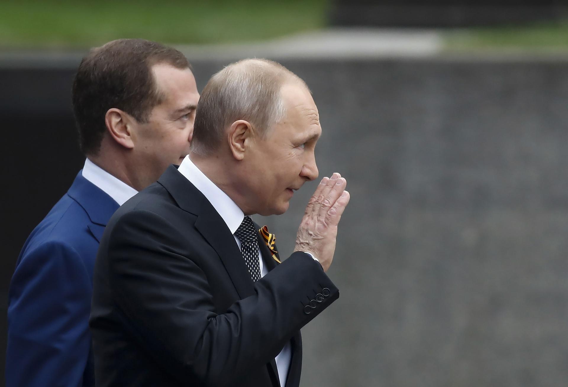 Russian President Vladimir Putin is shown waving with Prime Minister Dmitry Medvedev slightly behind him.