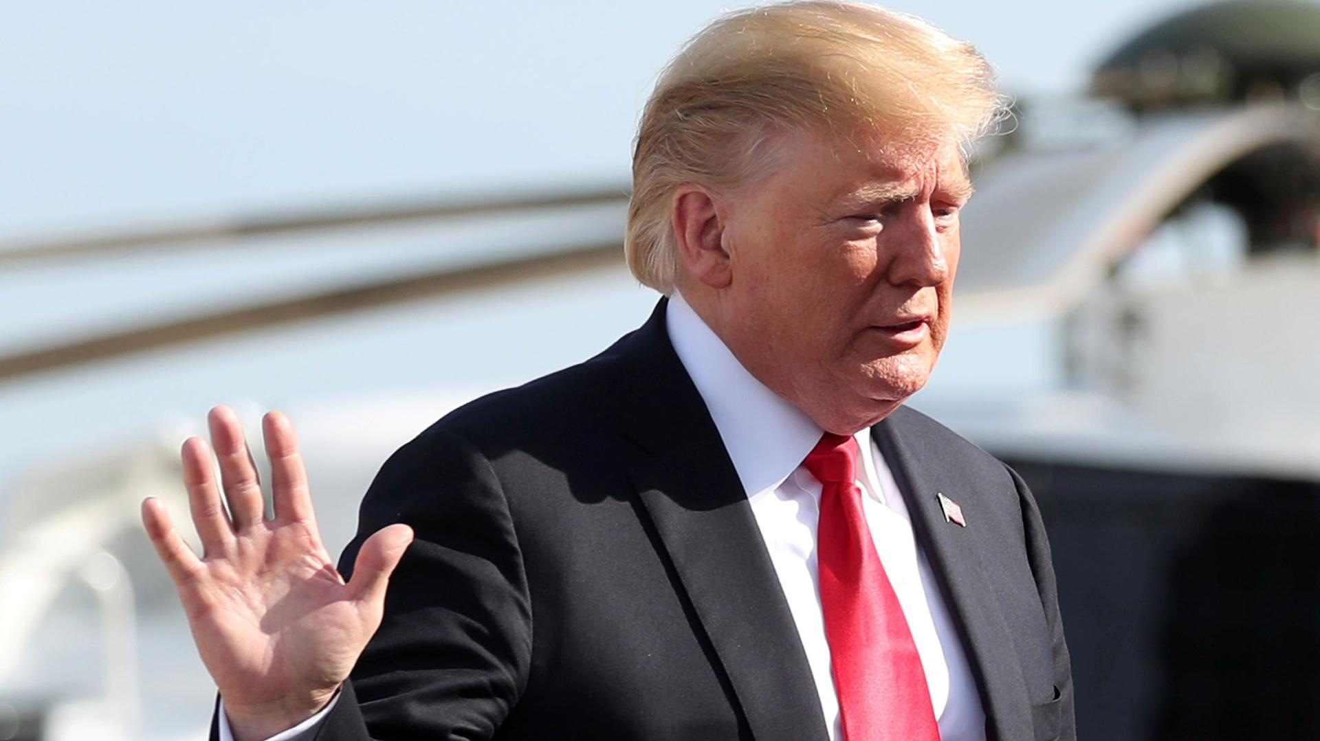 US President Donald Trump is shown with his right hand raised and wearing a dark suit with a red tie.