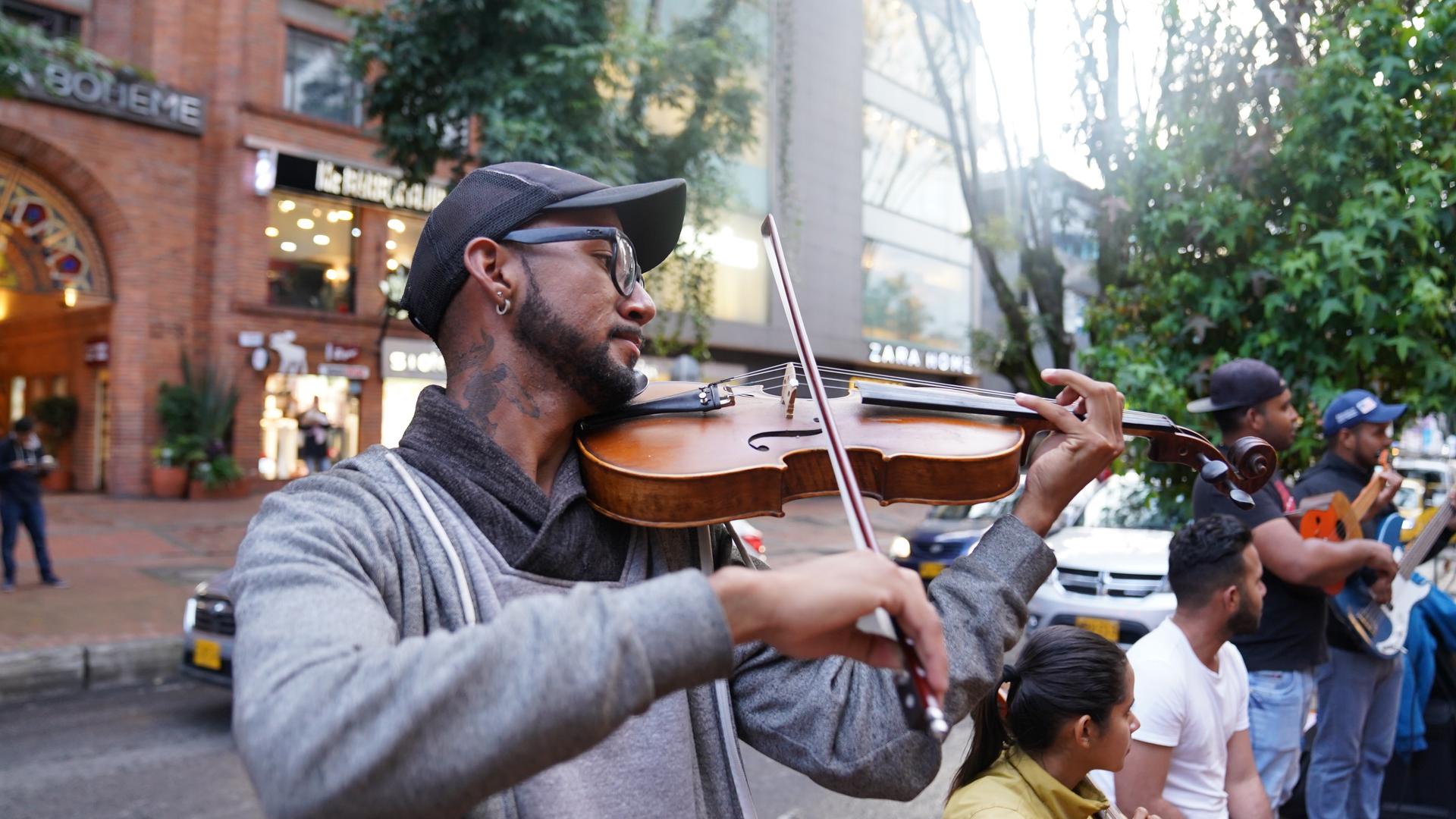 A young man with a neck tattoo and baseball cap plays violin on the street. 