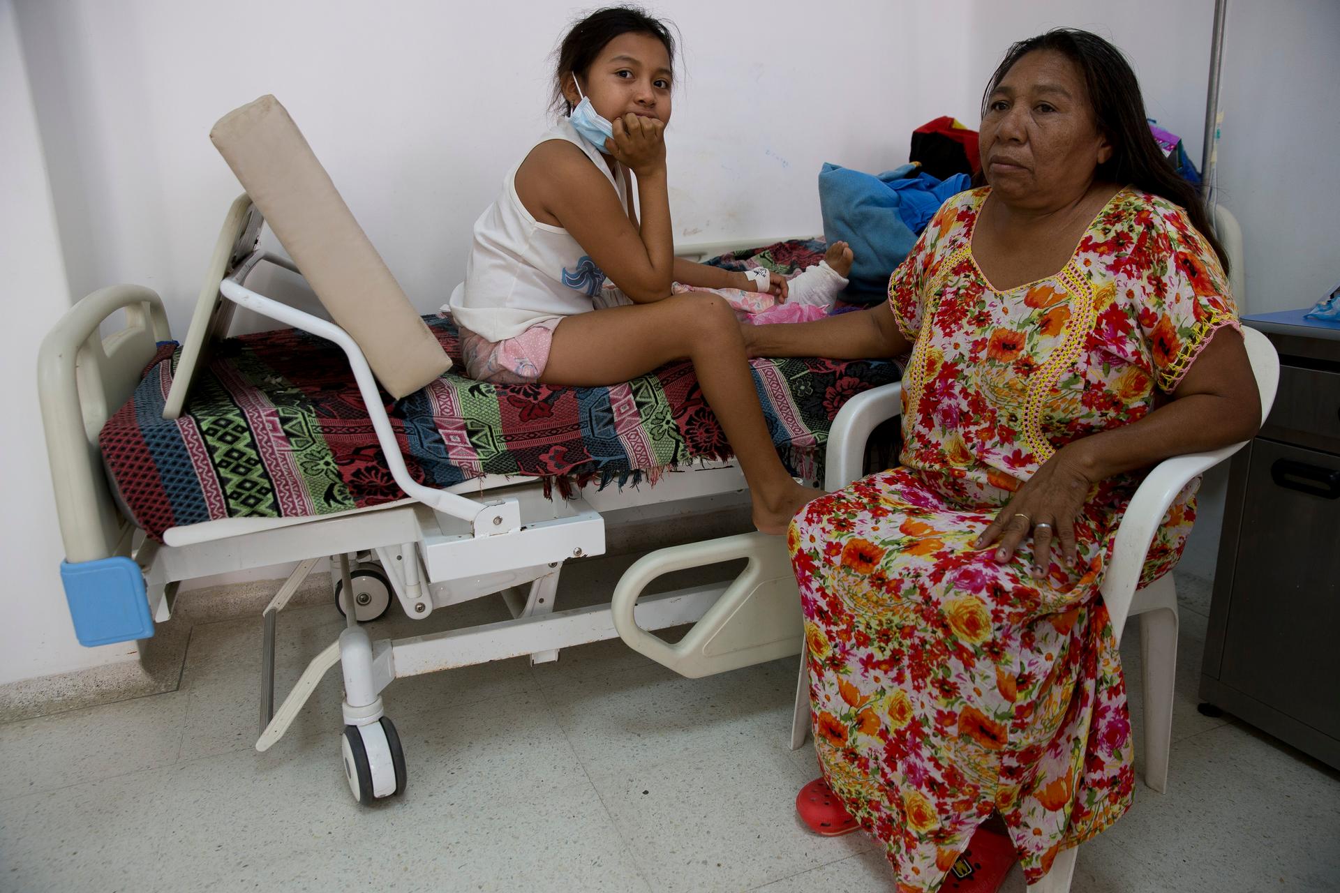 A young girl sits on a hospital bed with a fractured leg in bandages next to her mother.