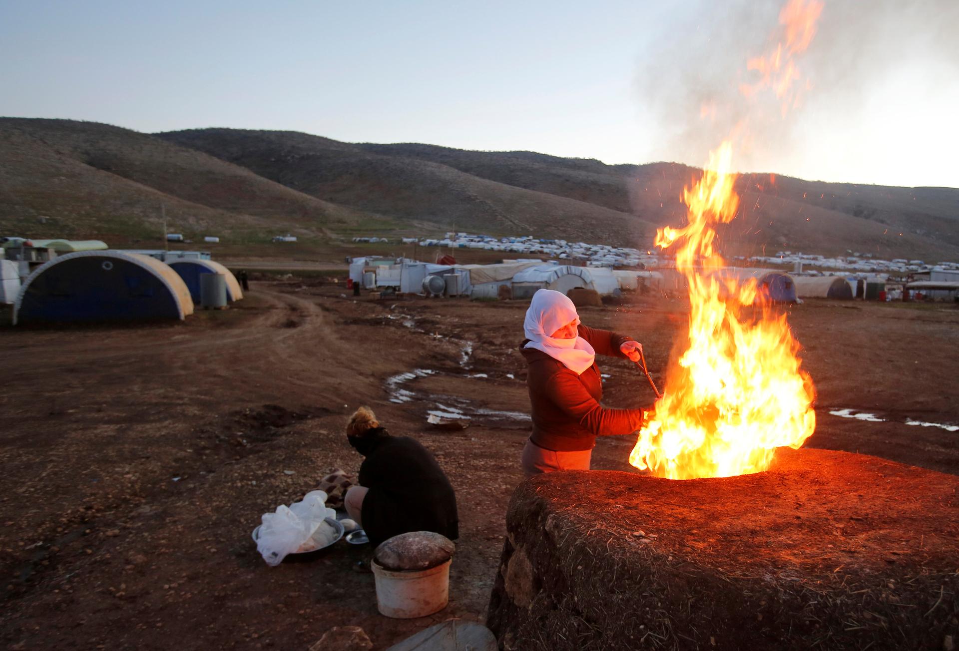 A woman prepares bread with huge fire at refugee camp near white tents.