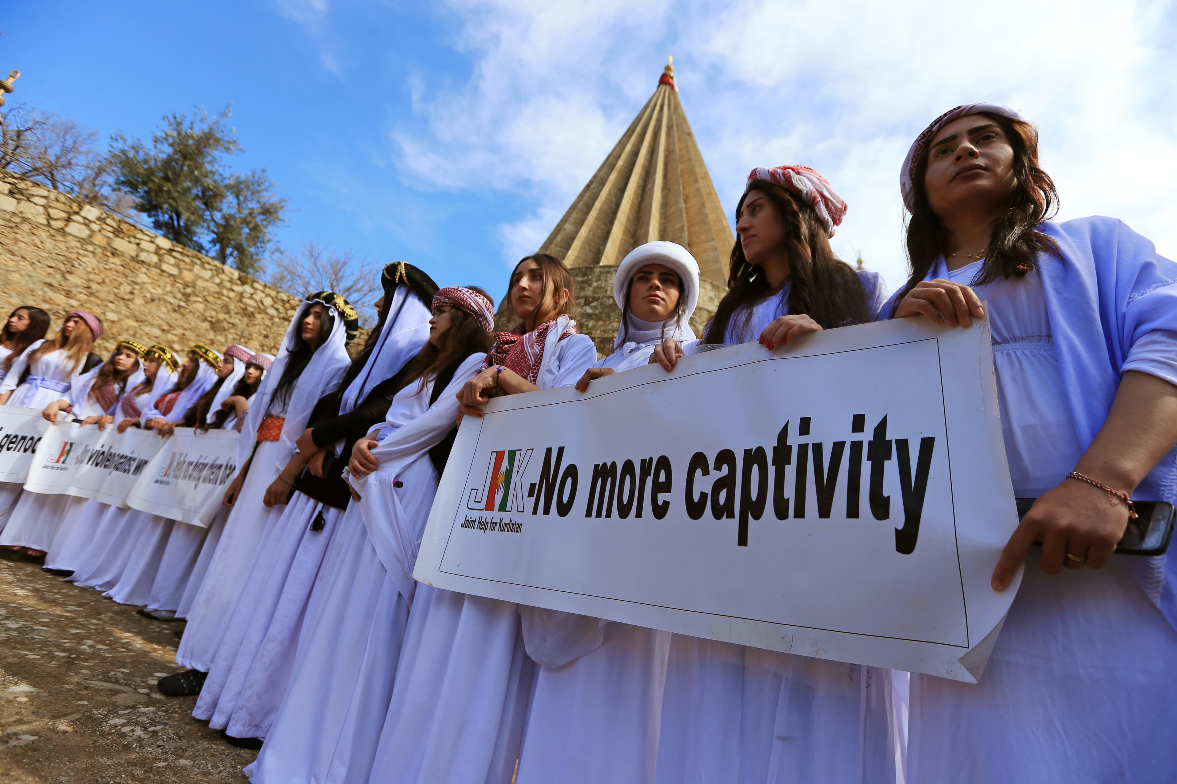 Women in white robes and turbans carry sign that says