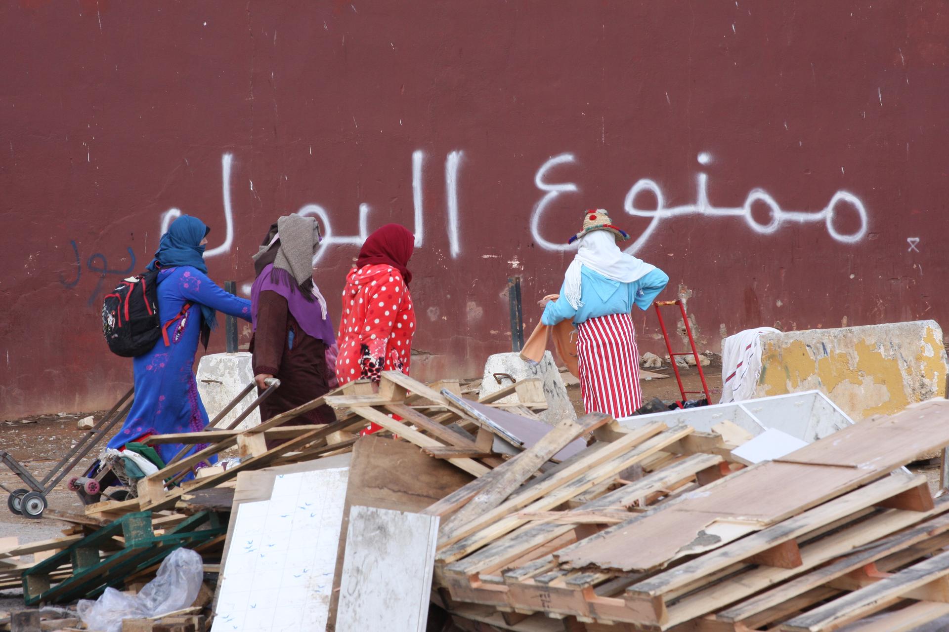 Women stand near pile of wooden debris against backdrop of red maroon wall with Arabic white letter graffitti.