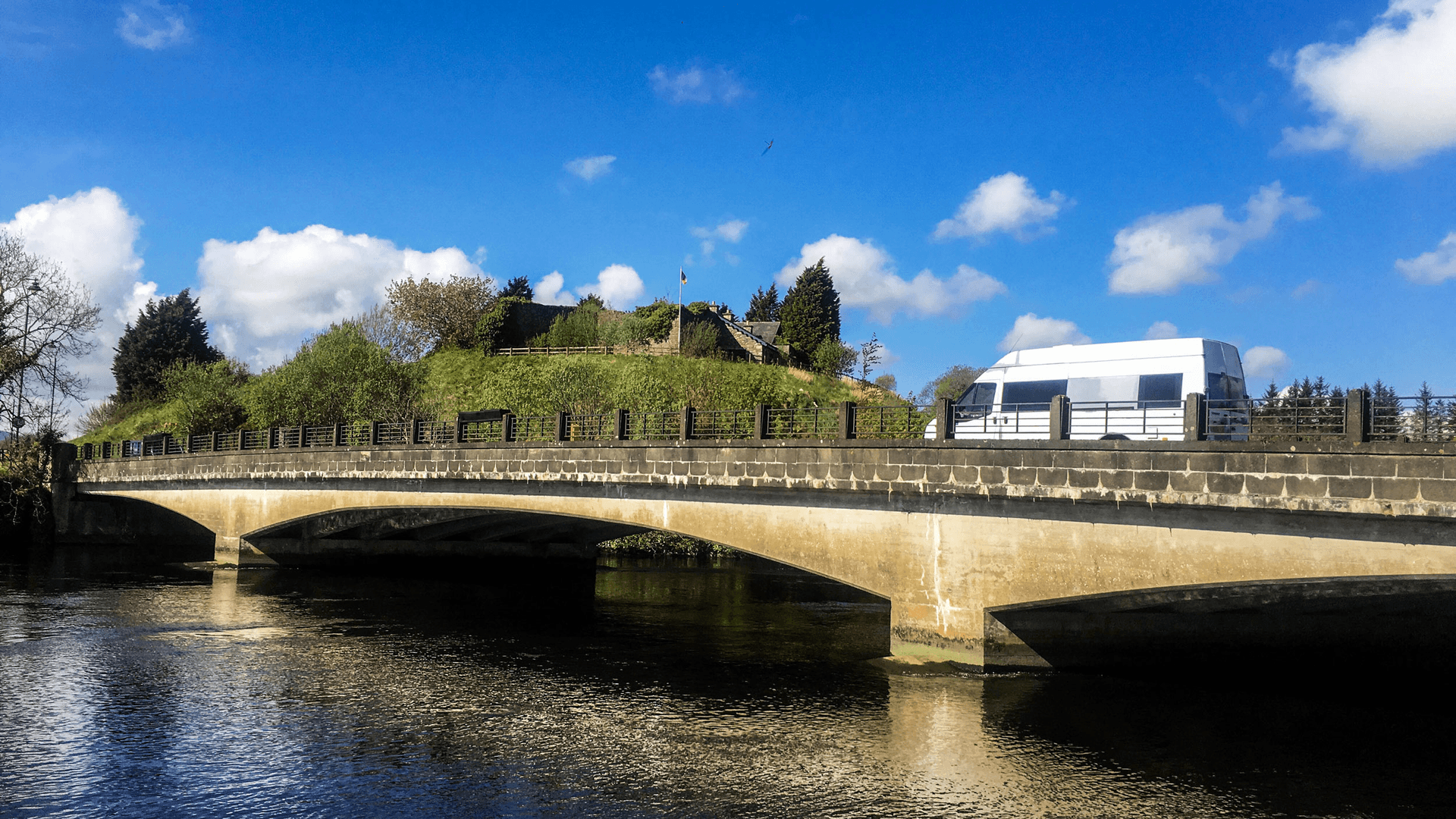 A white van travels over a grey stone bridge on a bright, sunny day under blue skies.  