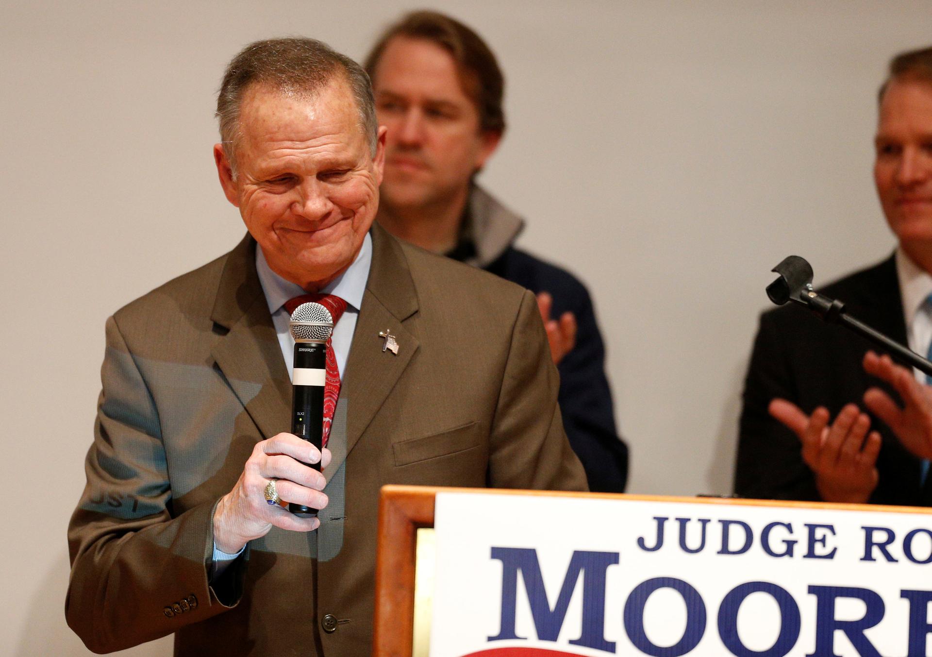 Republican US Senate candidate Roy Moore stands with microphone in hand behind a placard of his campaign poster.