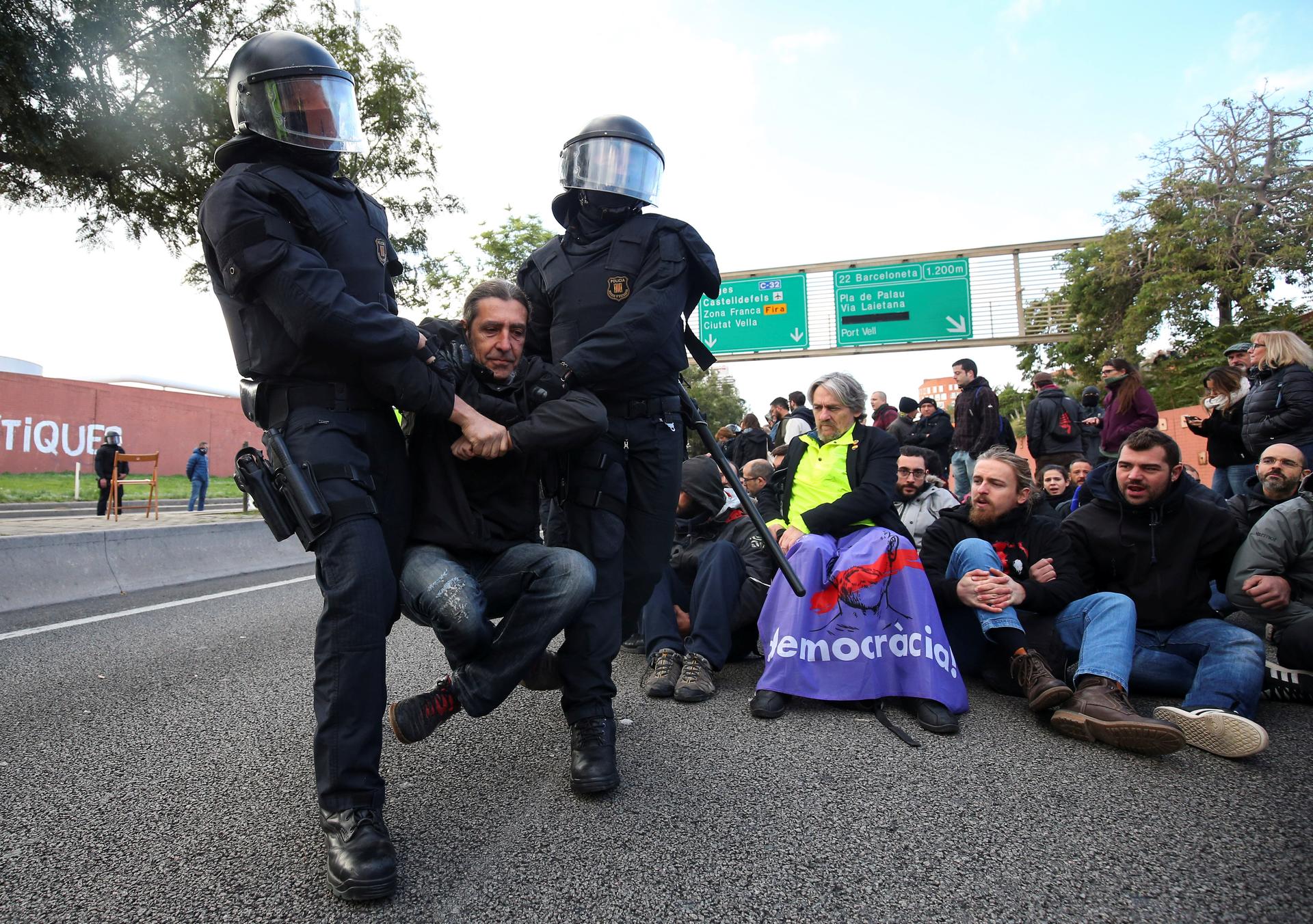 Two police officers wearing riot gear with face masks, move a man, cross-legged, from a road in Barcelona.