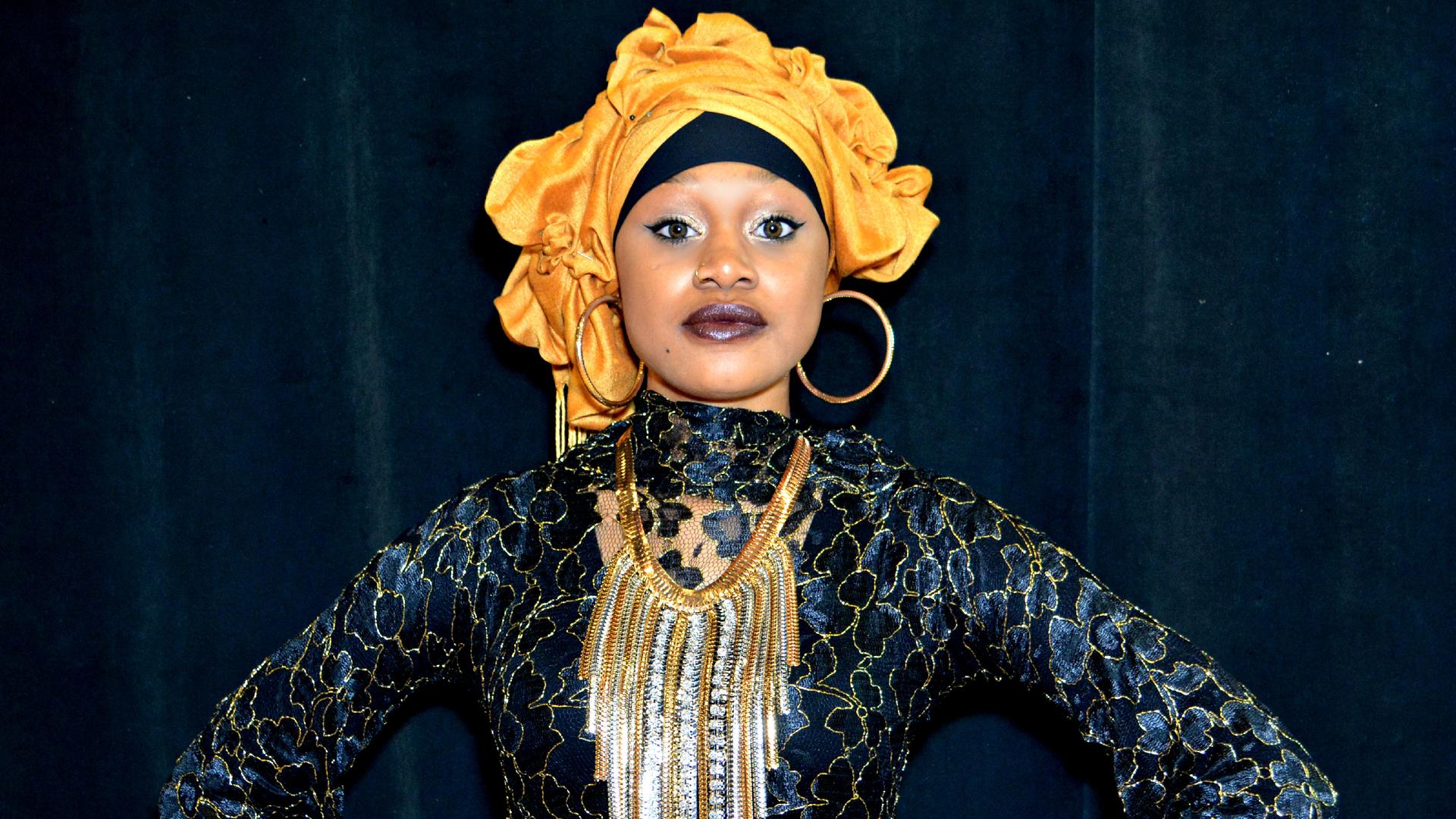 A woman wearing a headwrap and as shimmering blue and gold dress stands in front of a camera, with her hands to her hips. 