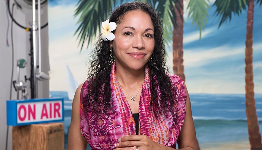 Woman in front of tropical set, "On Air" sign, with flower in her hair