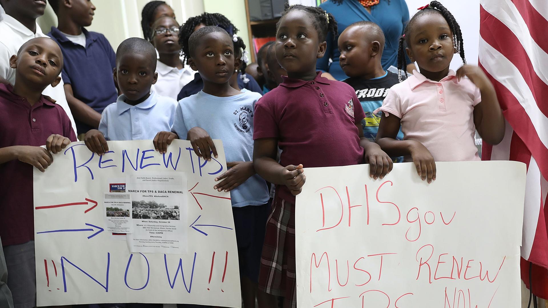 A group of several young children stand in a line with placards reading "Renew TPS Now."