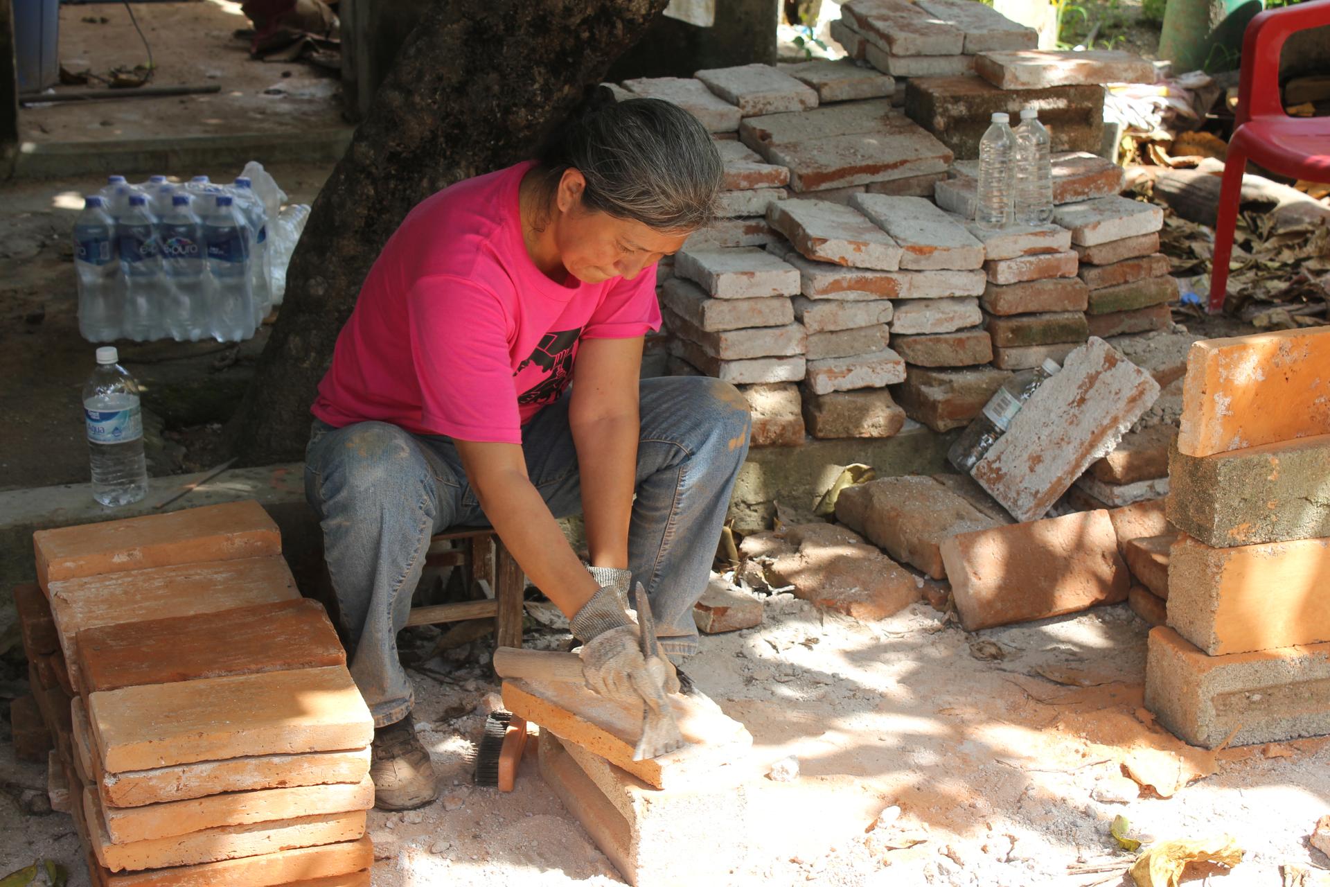 A woman scrapes mortar off of a salvaged brick with a flat end of a chisel so the brick can be reused.