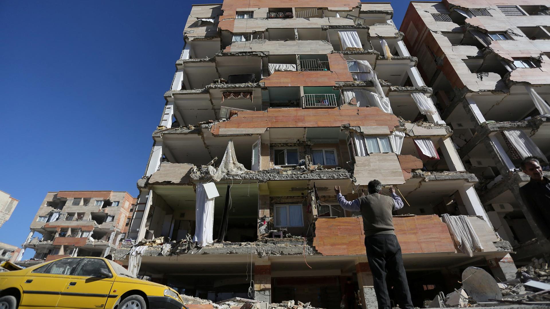 A man stands on the street with this arms raised looking up at a building with most of the walls broken off.