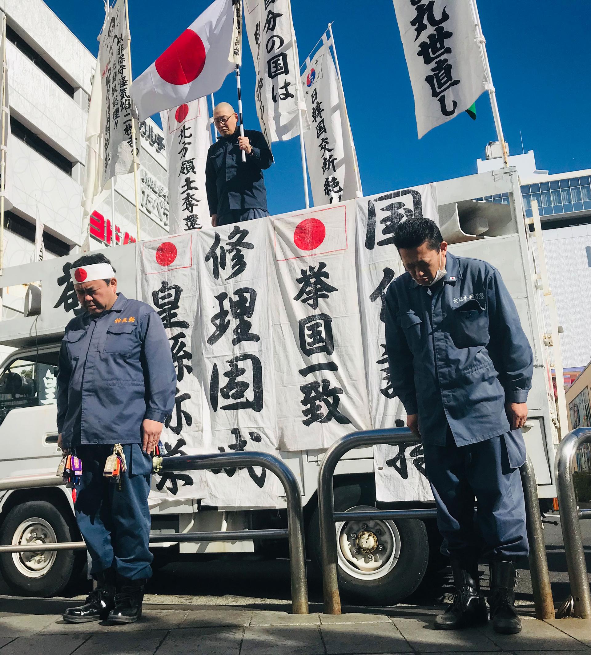 Men in blue jumpsuits stand at attention in front of a white van covered in banners written in Japanese and Japanese flags.