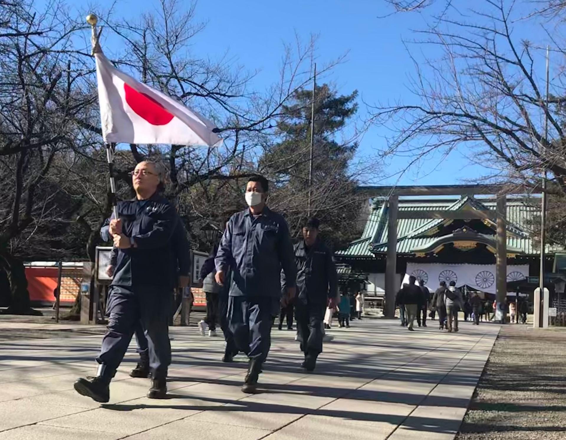 Men in blue jumpsuits march in a line across a plaza. The first in the line holds a Japanese flag above his head.