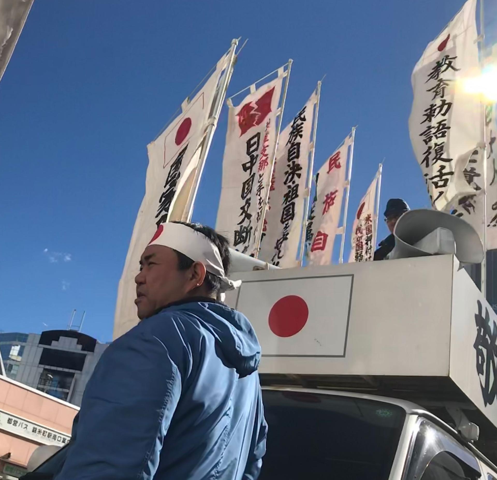 A man wears a bandana with the red circle from Japan's flag on his forehead. Behind him is a white van with Japanese banners and flags.