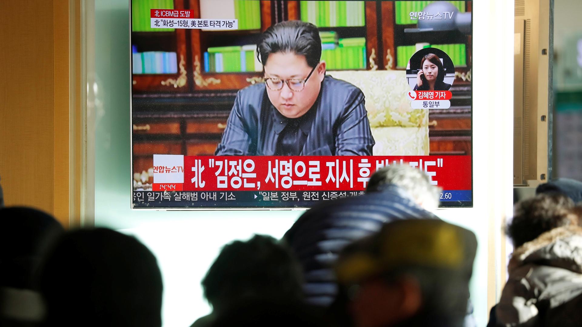 Several people out of focus are in the near ground with a TV monitor in the background showing Kim Jong-un.