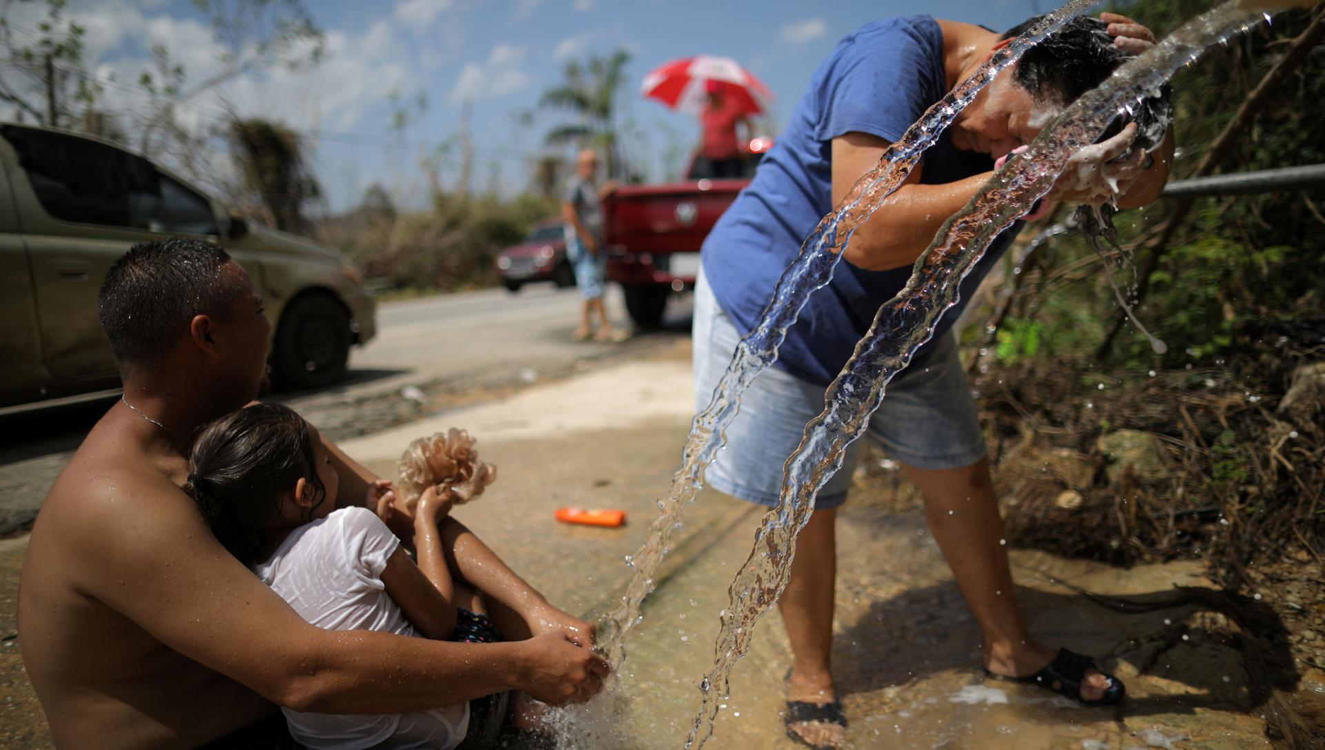 Water falls from a pipe onto the street. A woman in the background is bent at the waist and washes her hair in the running water, while a man holds a child and watches the woman. 
