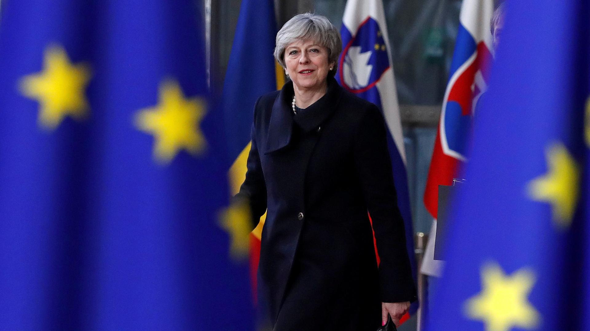 Britain's Prime Minister Theresa May wearing a dark blue jacket is picture between two EU flags in the foreground.