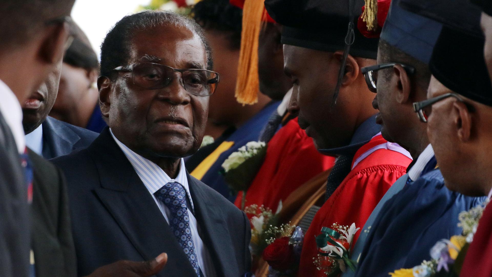 Zimbabwe President Robert Mugabe stands center with a blue suit and tie shaking the hands of college graduates.